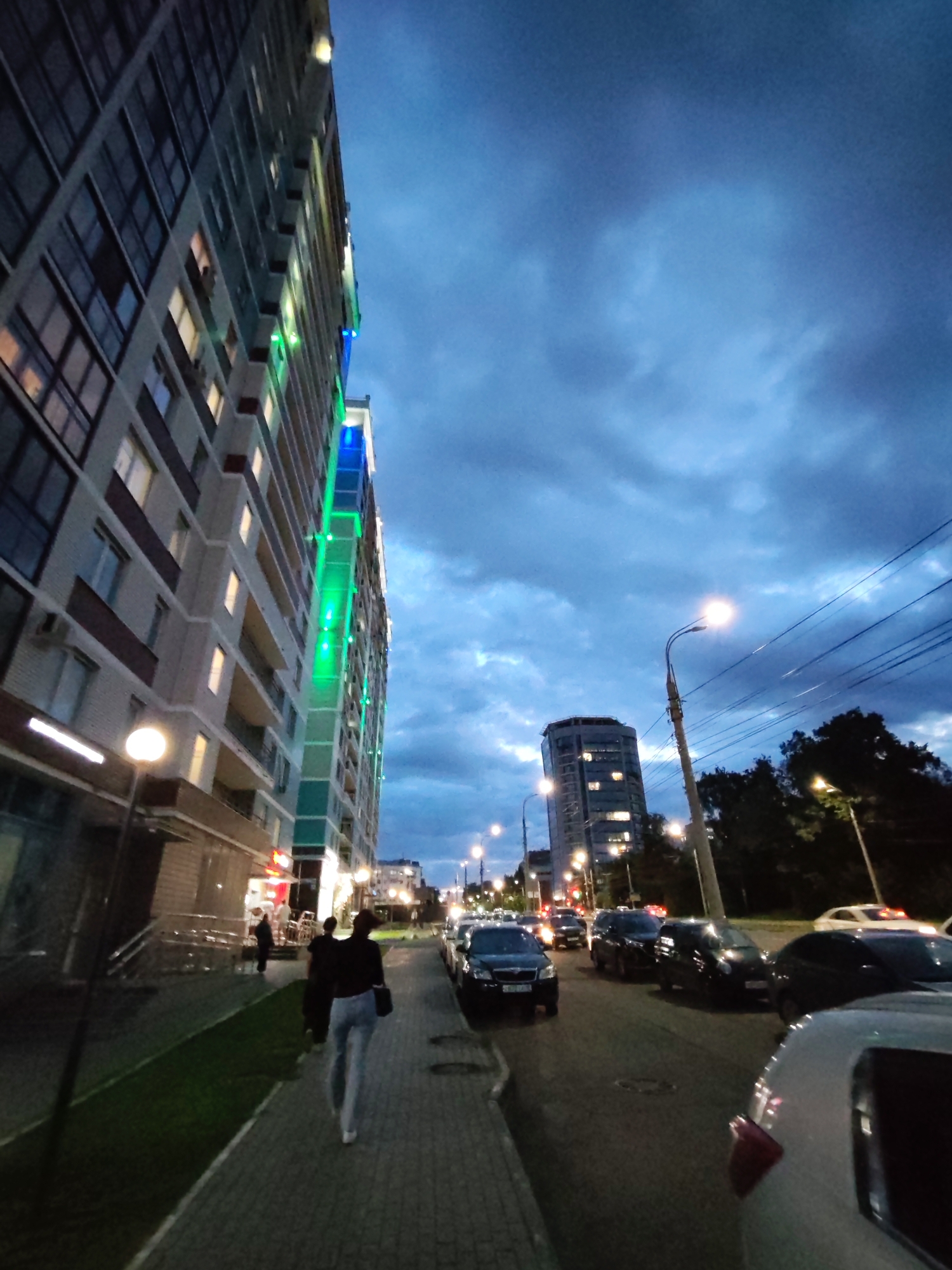 The image depicts a street scene at dusk in Izhevsk, Russia, with a tall, modern apartment building as the dominant structure on the left. The building features a grey facade, balconies, and vertical strips of green and blue lighting. A sidewalk runs alongside the building, with a few people walking on it. Across the street, a row of parked cars is visible, along with streetlights and a taller, cylindrical building in the distance. The sky is cloudy and dark blue, indicating the transition from day to night. The overall atmosphere is urban and contemporary.