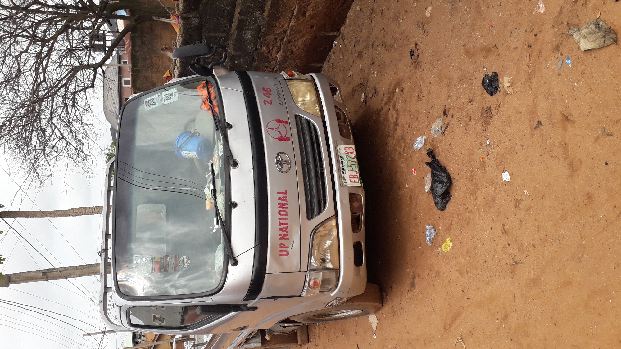 The image depicts a silver Toyota Dyna truck parked on a dirt road in Nkpor, Nigeria. The truck has the lettering "UP NATIONAL" and "246" on the front. The license plate reads "EBJ-572XB". The ground is littered with trash, including plastic bags and wrappers. There are trees and a building visible in the background. The sky is overcast.
