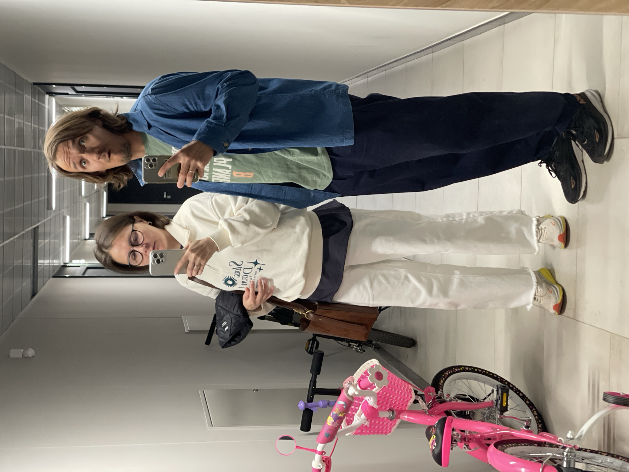 Two people, a man and a woman, are taking a selfie in a hallway. The man is wearing a blue shirt over a green t-shirt and navy pants, with black sneakers. The woman is wearing a white sweatshirt with a design, white pants, and tan sneakers. She also wears glasses. They are holding phones up to take the picture. Behind them, there's a pink child's bicycle, an adult bicycle, a black backpack, and a brown bag. The hallway has white walls and a light-colored floor. The location is Unknown, Unknown.