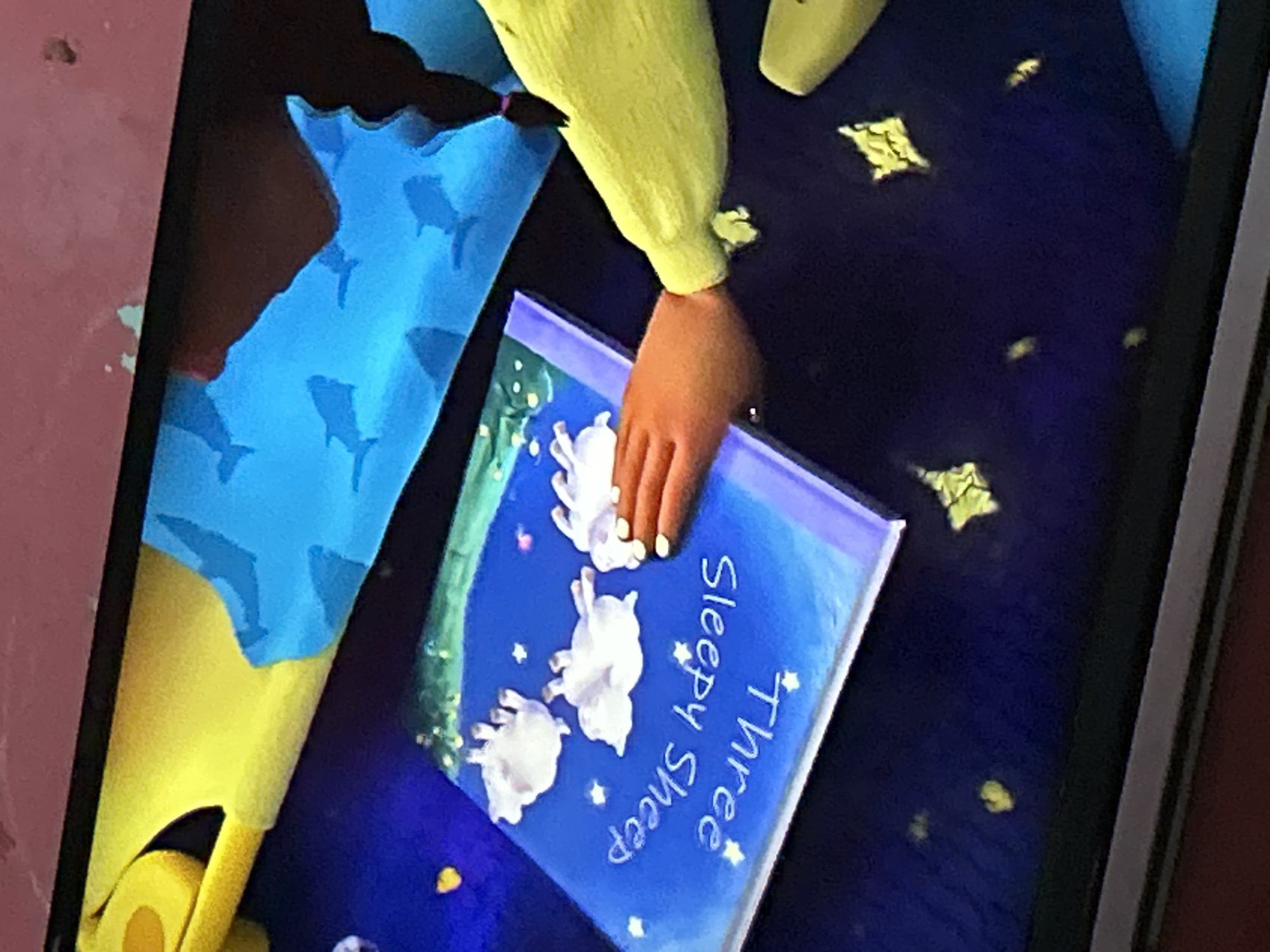 The image shows a close-up view of a screen displaying a book cover with the title "Three Sleepy Sheep." Three white sheep are depicted upside down on the blue cover, which also features scattered yellow stars. A person's hand, with light skin and visible fingernails, is seen touching the book cover. The background of the screen shows a blue surface with dark silhouettes of sharks. To the left of the screen, a yellow object with a rounded edge is partially visible. The overall scene suggests an interactive display or a digital presentation. The question asks for the location of this scene in Depok, Indonesia, however, there is no information within the image to determine its geographical location.