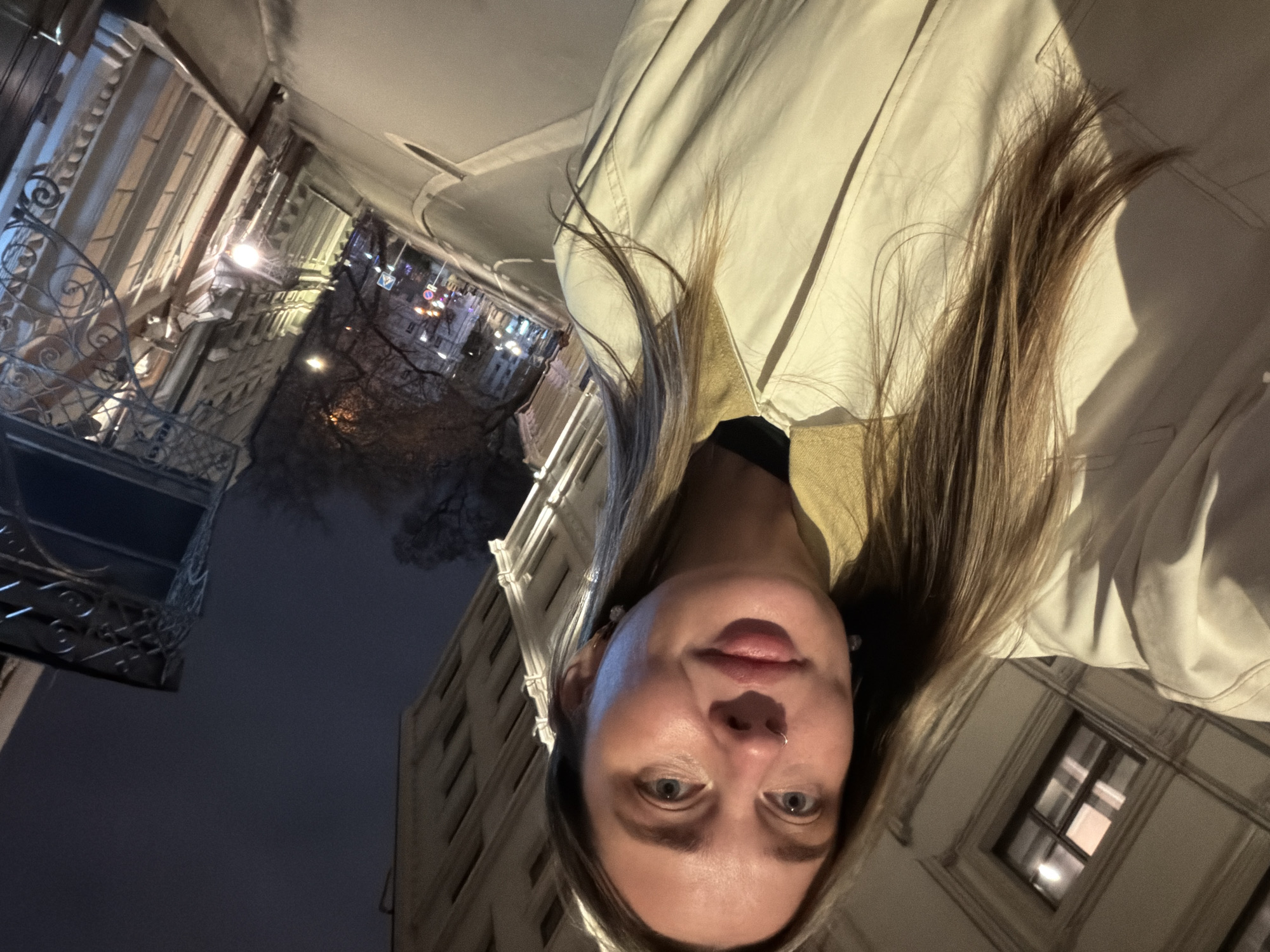 The image depicts a woman wearing a light-colored trench coat and standing in an urban environment. She is looking directly at the camera. The background shows buildings illuminated at night. The location appears to be Moscow, Russia, indicated by the architectural style of the buildings and street lighting.
