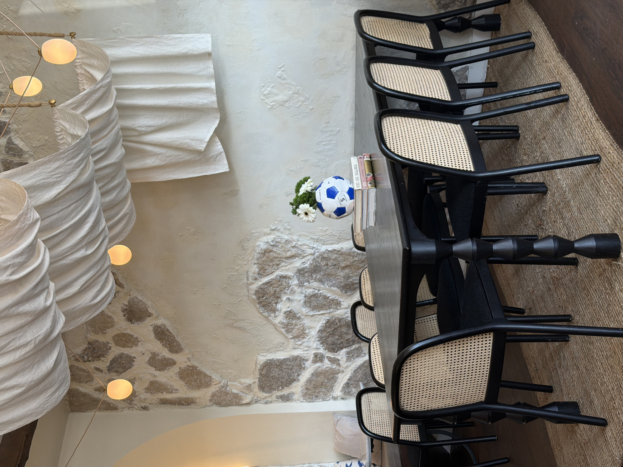 The image depicts a dining area with a rustic and modern aesthetic. The scene is dominated by a long, dark wooden table surrounded by black chairs with woven seats. On the table, a blue and white soccer ball sits next to a small bouquet of white flowers and a few books. The background features a textured white wall with exposed stone elements and large, draped white fabric installations that resemble lampshades. Several light fixtures, with round, glowing bulbs, hang from above. The floor is covered with a natural fiber rug. The lighting suggests it might be daytime or evening, with the warm glow of the lights creating an inviting atmosphere. The overall impression is one of casual elegance, possibly in a restaurant or a well-decorated home. There is no visible text in the image.