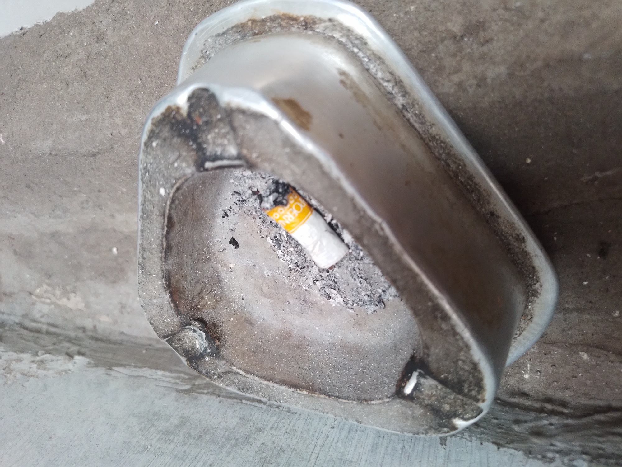 The image shows an outdoor metal ashtray filled with cigarette butts and ash. The ashtray appears worn and tarnished. It is situated on a concrete surface, possibly a sidewalk or ledge, with a textured, grey wall in the background. The overall scene suggests a public space in Batu, Indonesia, where smoking is permitted.