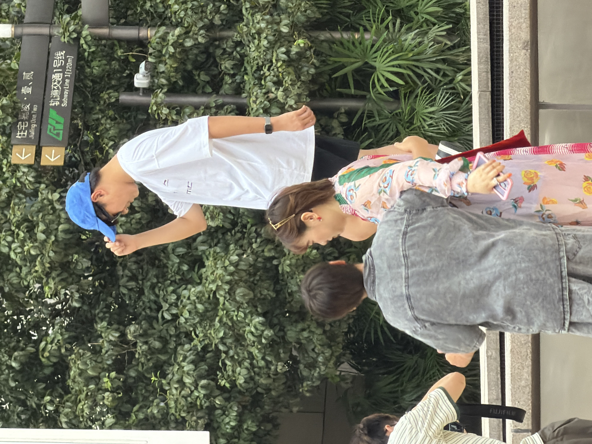 In Yuzhong District, China, a medium shot captures several individuals against a backdrop of lush green foliage and a grey stone wall. A young man wearing a blue baseball cap and a white t-shirt stands prominently, adjusting his cap. To his right, a signpost indicates directions with Chinese characters and English translations. Partially visible are two other individuals: one in a grey denim jacket and the other in a floral patterned dress holding a pink smartphone. Another person, wearing a striped shirt, is also visible on the left. The scene appears to be outdoors, possibly a public area or a walkway.