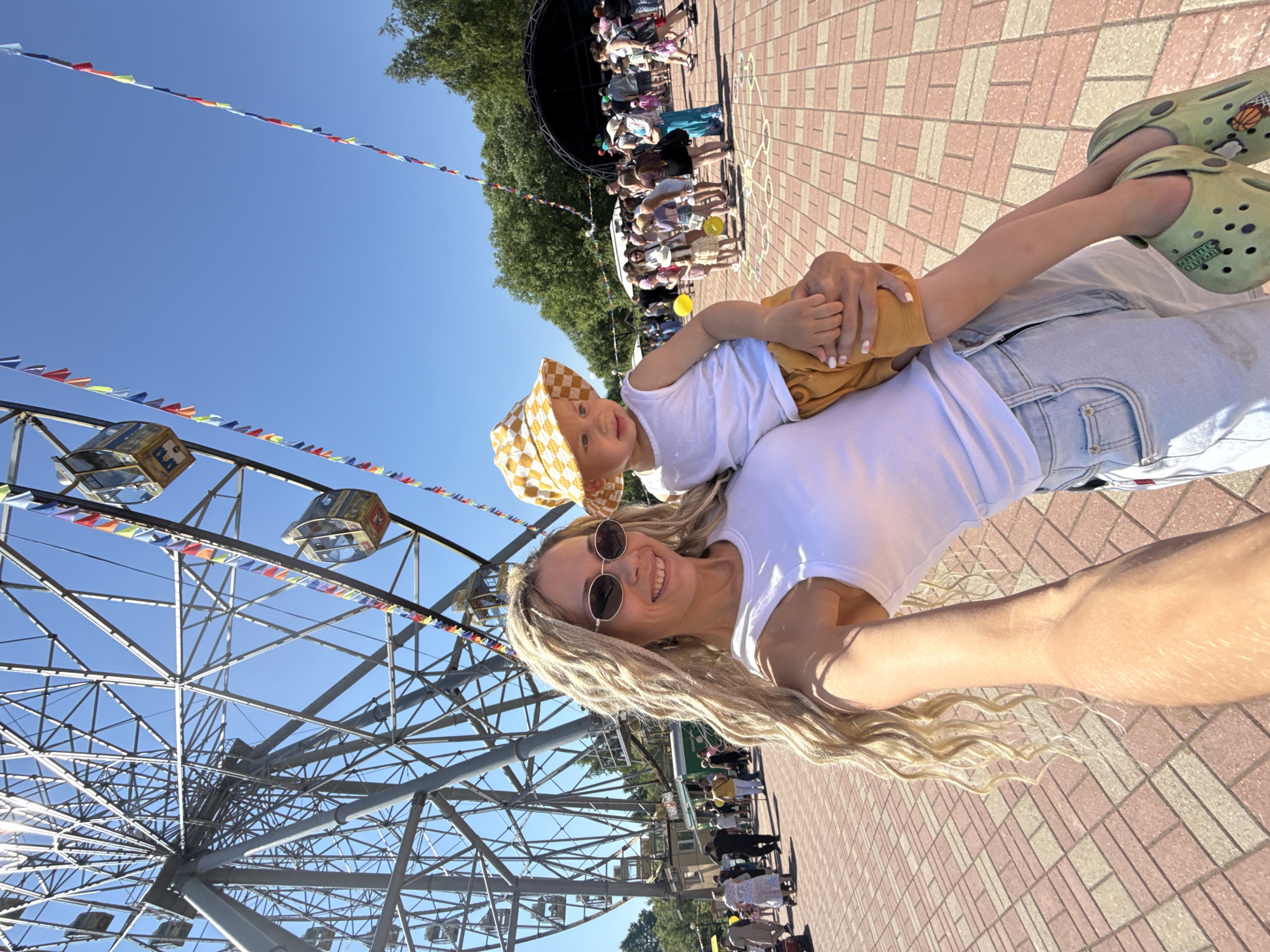 In an outdoor amusement park setting, a woman with blonde dreadlocks and sunglasses, wearing a white tank top and light-wash jeans, holds a young child. The child, wearing a checkered bucket hat and a white tank top with yellow shorts, is held in her arms. They are positioned in the foreground, with a large Ferris wheel visible in the background. The ground is paved with a pink and tan brick pattern. Numerous people are scattered throughout the background, some appearing to be in a queue for attractions or browsing. String lights with colorful flags are suspended overhead. The sky is a clear blue. The scene appears to be illuminated by bright daylight.