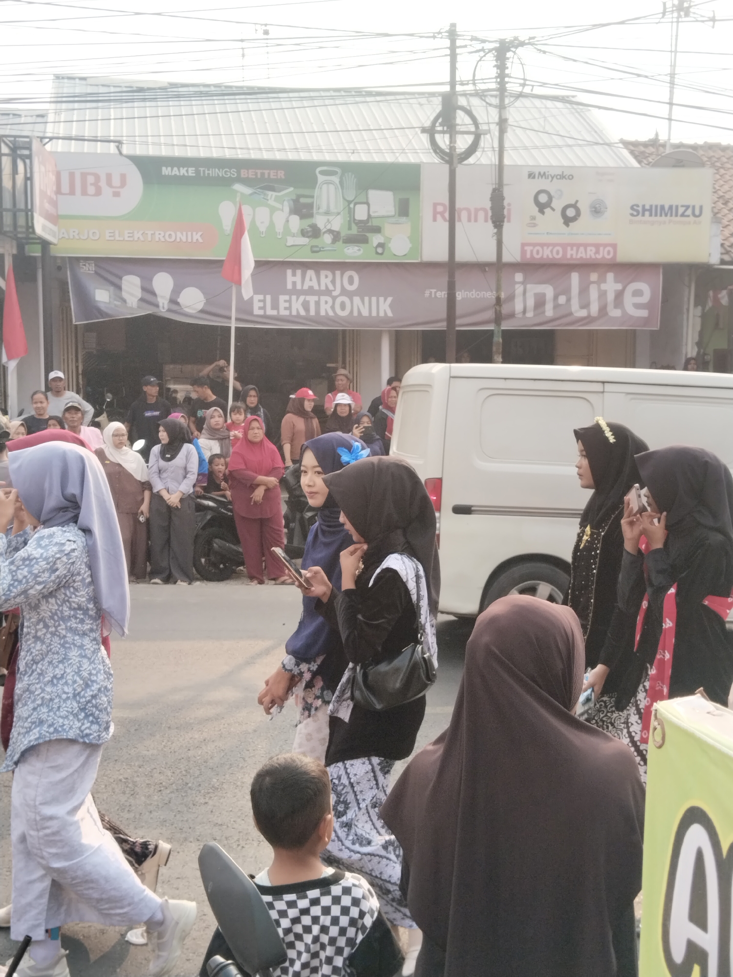 In Balapulang Kulon, Indonesia, on a street, a crowd of people gathers in front of a building with multiple signs. One prominent sign reads "UBY MAKE THINGS BETTER HARJO ELEKTRONIK". Another sign features the brand names "Miyako", "Rinnai", and "SHIMIZU". Below these, "TOKO HARJO" and "in-lite" are visible. A white van is parked on the street. Several individuals are walking, including women in hijabs and traditional attire. Some are holding phones, suggesting they might be taking pictures or videos. A young boy in a checkered shirt is seated, facing away from the camera. Two Indonesian flags, one red and white, are displayed. The environment appears to be an outdoor commercial area with overhead wires and power poles.