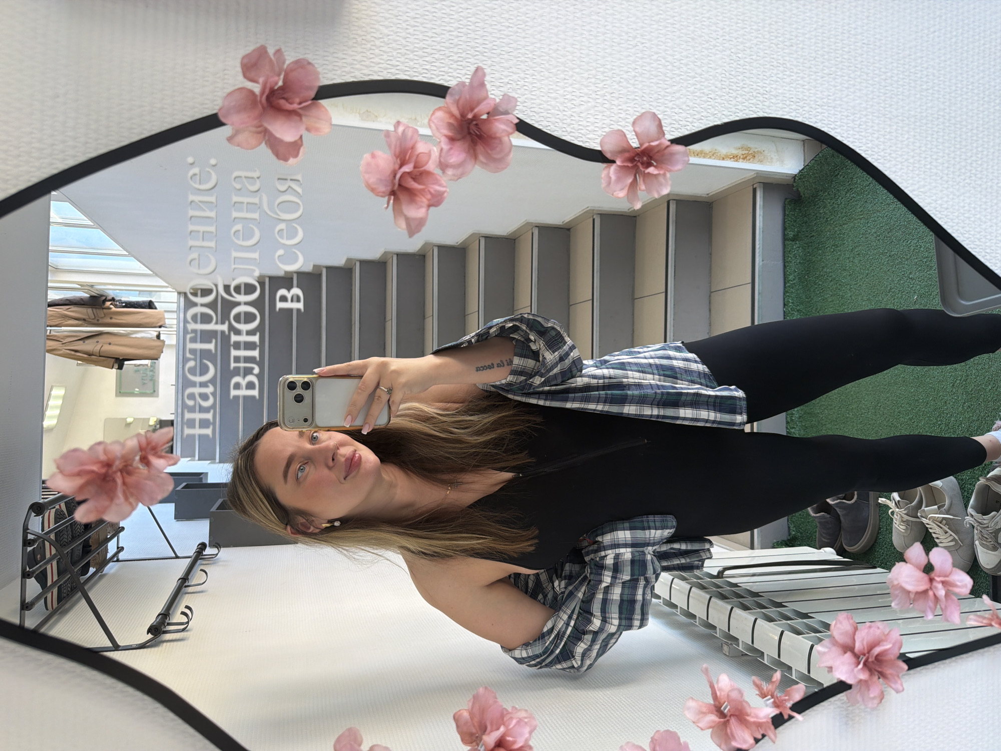 A woman is posing in front of a mirror, taking a selfie with her phone. She is wearing a black top, black leggings, and a blue and green plaid shirt. The mirror is decorated with pink flower stickers. The reflection shows a staircase and a green patch of flooring. In the background, there's a coat rack. The writing on the mirror reads "Настроение: влюблена в себя". Based on the location information provided, this scene is located in Chaikovsky, Russia.