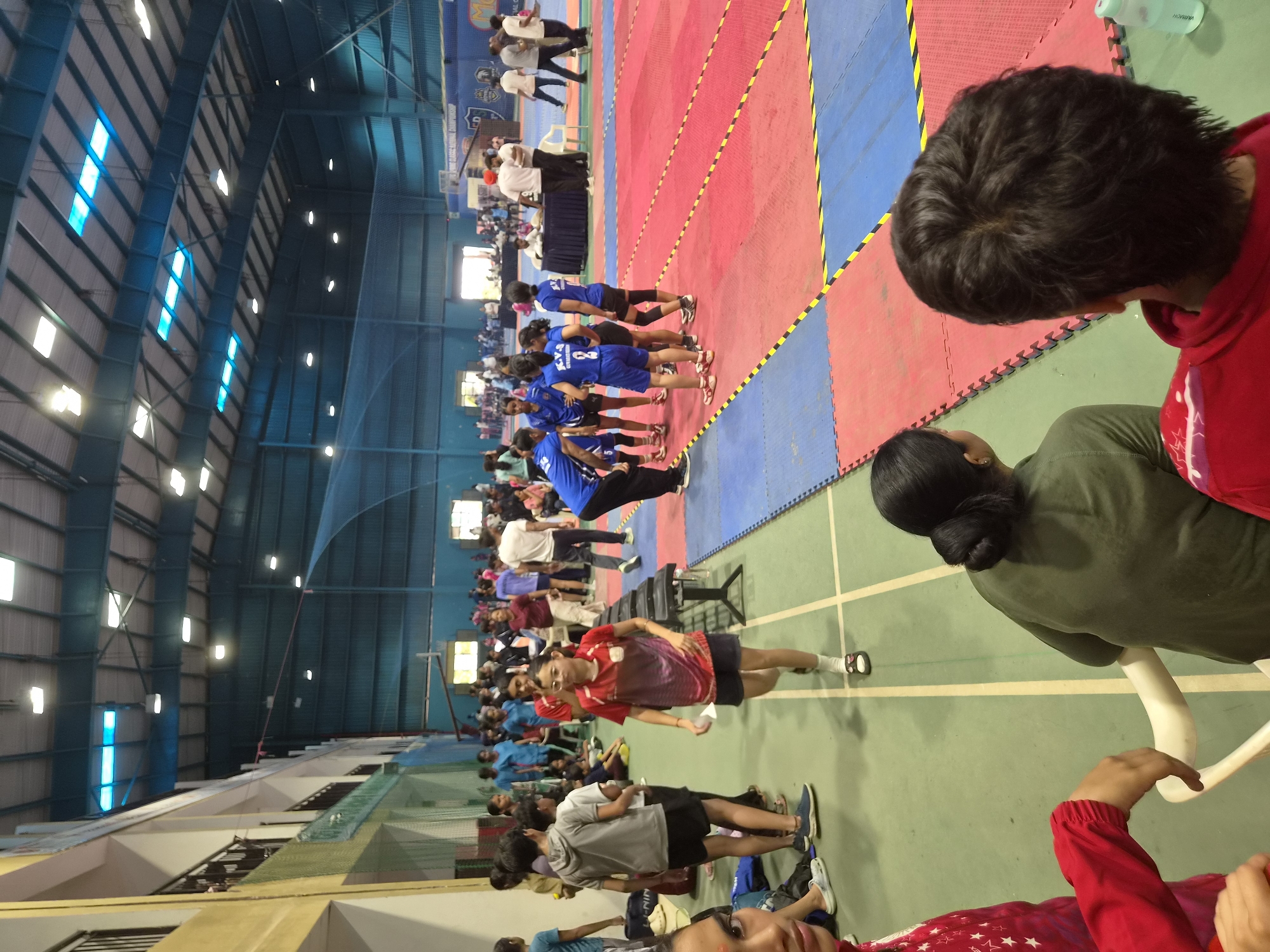 This media file depicts an indoor sports event, likely a kabaddi match or tournament, taking place in Phagwara, India. The scene is a large gymnasium with a high ceiling and a distinctly marked court, divided into red and blue sections. Rows of people, seemingly participants and spectators, are visible on the court and along the sides. Many individuals are wearing athletic attire, predominantly blue and red.
In the foreground, two individuals are captured from the back, one wearing a red hooded sweatshirt and the other a green athletic top. Their presence suggests they are part of the event, either as participants waiting for their turn or as observers. The general atmosphere appears to be one of an organized sporting event, with participants lined up and ready.
The background shows the structure of the gymnasium, with large windows and lighting fixtures on the ceiling. The presence of a large net and other sports equipment is also implied by the setting. The red and blue mats on the court, along with the white lines marking the boundaries, are characteristic of kabaddi courts.
The image is taken during the daytime, evidenced by the natural light filtering through the windows. The focus is primarily on the people and the sports arena, with no specific indication of weather conditions. The overall mood seems to be energetic and competitive, typical of a sporting event. No discernible text is visible in the image, other than what might be on clothing or signage in the distance, which is illegible.