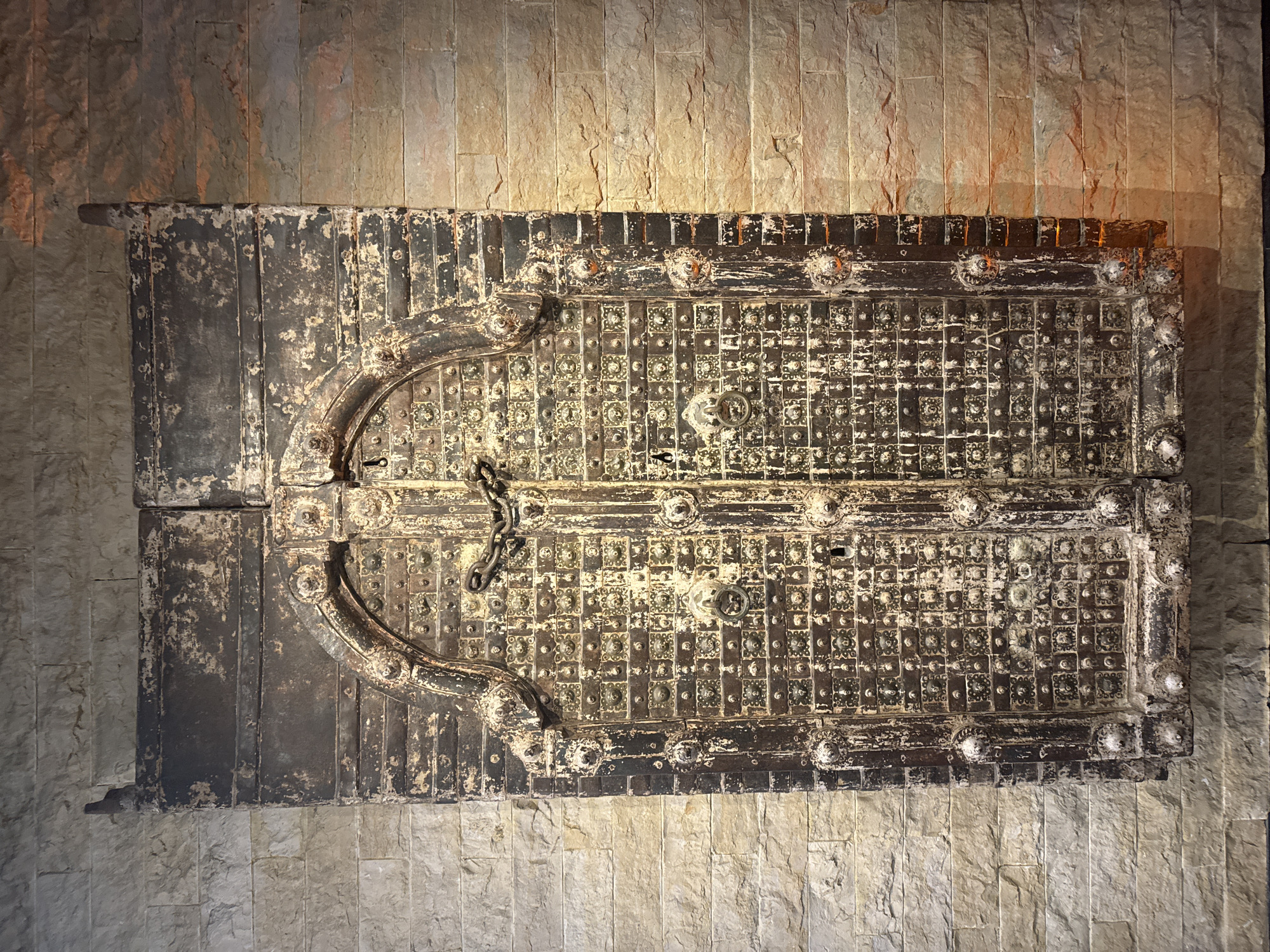 The image shows an old, ornately decorated, double-door structure set against a stone wall. The doors appear to be made of dark metal or wood with metal reinforcement, possibly iron. They have a patterned design with small square panels and decorative rosettes. There is a central latch with a chain. The structure looks like a medieval or antique chest or storage box. The stone wall is made of rectangular blocks of varying shades of beige and grey. There is a light source above, casting a warm glow. The city, Noordwijk, Netherlands, is not evident in the image.