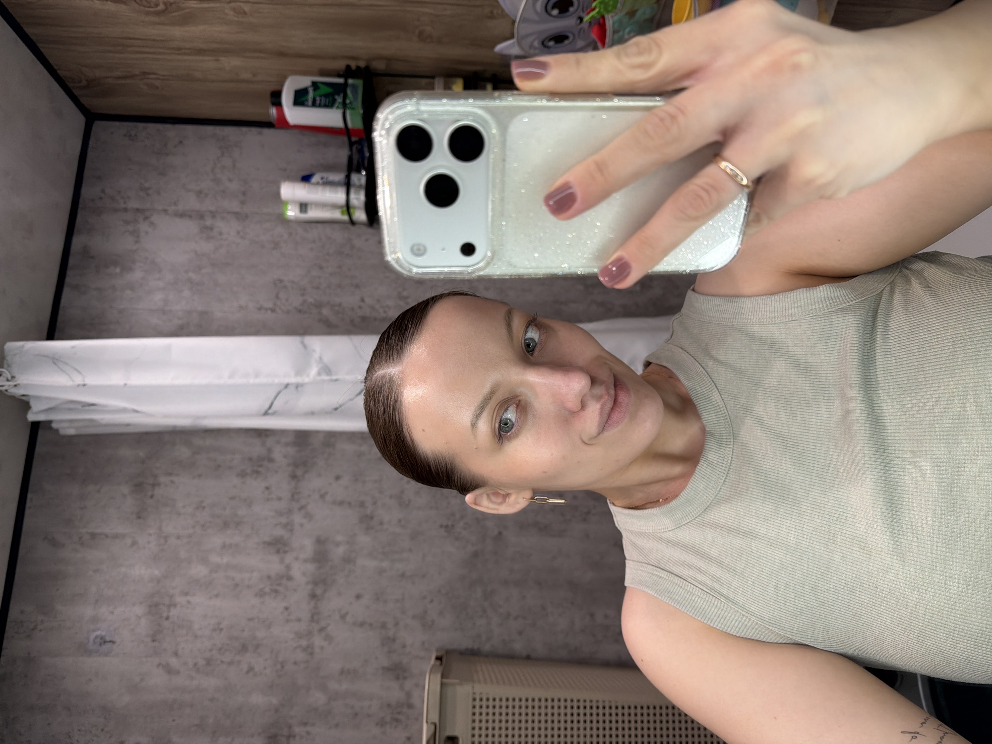 This image captures a woman taking a selfie in what appears to be a bathroom setting in Reshetnikovo, Russia. The woman, with her hair pulled back and striking light-colored eyes, looks directly at the camera with a calm, subtle smile. She is wearing a light sage or greenish-grey ribbed sleeveless top. Accessories include a gold-toned ring on her right ring finger and a modern, elongated gold-toned earring in her left ear. Her fingernails are painted a muted purplish-brown.
She holds a smartphone, likely an iPhone due to its triple camera array, encased in a clear, sparkling glitter case. The background reveals elements typical of a bathroom: a white and grey marble-patterned shower curtain or liner on the left, and grey, textured wall panels or tiles. Above, a wooden shelf or cabinet edge is visible, holding various toiletries. Among these bottles, some green and white, partially visible text includes "DOVE" and "SHAMPOO." These items are secured by black ties. A small, grey, cartoon-like figurine resembling Grogu (Baby Yoda) is perched on the wooden shelf. A light-colored, possibly beige, grated object, perhaps a laundry hamper or heater, is partly visible in the lower right corner. The scene is lit by indoor lighting, with no specific time of day or weather discernible.