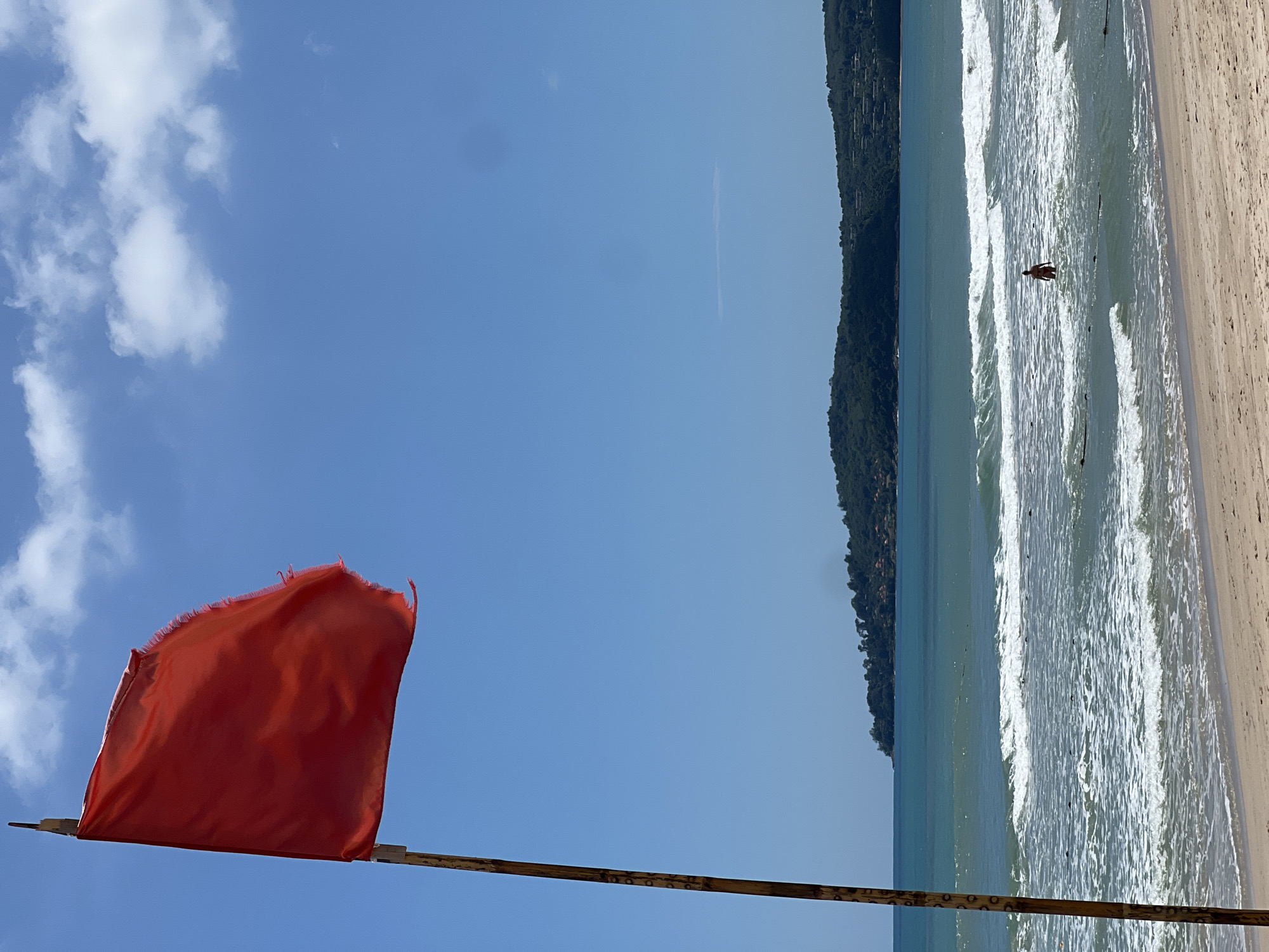 A red flag waves from a wooden pole on a sandy beach. The flag, appearing tattered at the edges, is positioned in the left foreground of the image. Beyond the beach, the turquoise ocean stretches out with moderate waves breaking and creating white foam. In the middle ground, a lone person is standing in the water. In the background, a lush green, tree-covered island with scattered buildings rises from the sea. The sky above is a clear, vibrant blue with scattered white clouds.