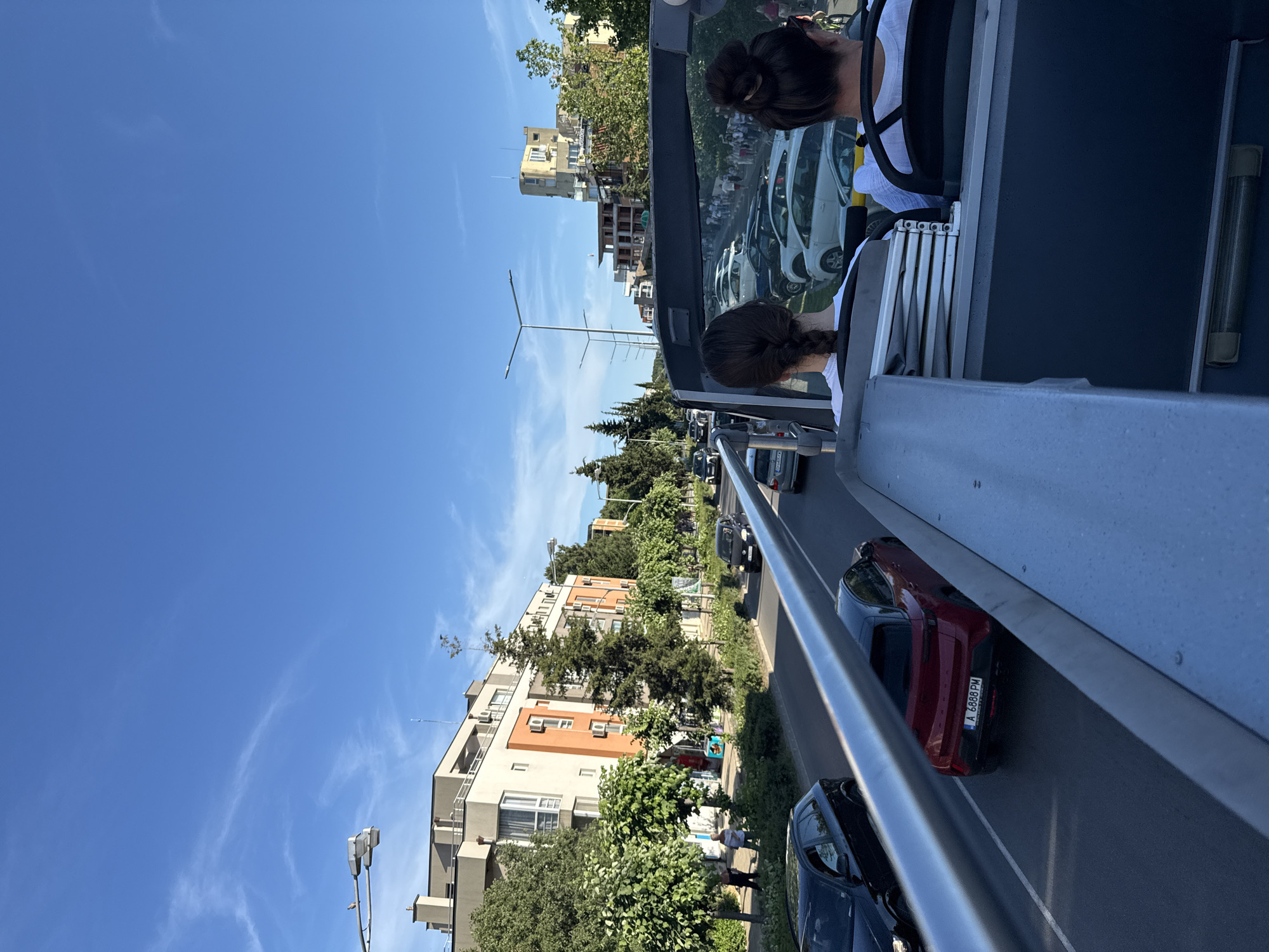The image captures a street scene in Nesebar, Bulgaria, viewed from the upper deck of a double-decker bus. The bus is traveling on a road with multiple lanes of traffic, bordered by buildings and trees. Two passengers, both women with dark hair and wearing white tops, are seated in the foreground, facing away from the camera. The woman on the left has her hair in a braid, and the woman on the right has her hair in a bun.
The street is lined with modern buildings, some with orange and beige facades, and several mature trees. Cars are visible in traffic, including a black SUV on the left, a red hatchback with Bulgarian license plates "A 6888 PM" in the middle lane, and other vehicles further down the road. The sky is clear blue with some wispy clouds. In the distance, more buildings and trees are visible, suggesting an urban or suburban environment. The overall impression is of a sunny day in a bustling city.