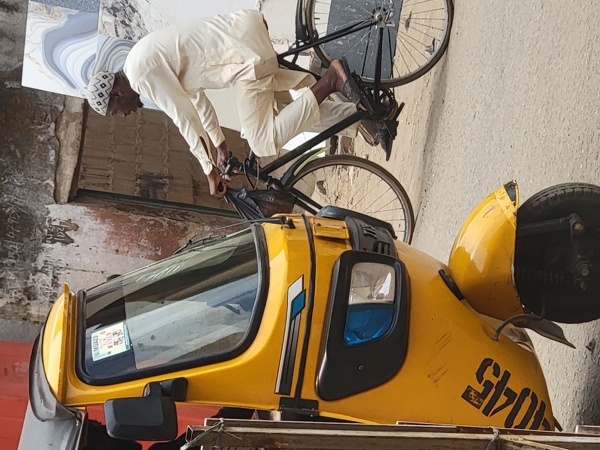 A man in a white robe and patterned cap is straddling a black bicycle, leaning forward to adjust something at the front, likely on a street in Maiburiji, Nigeria, as evidenced by the "U MUAZU" vehicle permit sticker on the windshield of a yellow three-wheeled taxi or rickshaw in the foreground. The vehicle has black accents and the number "6170" is painted on the side. Part of a building with patterned tiles can be seen behind the man. The background suggests an urban environment.
