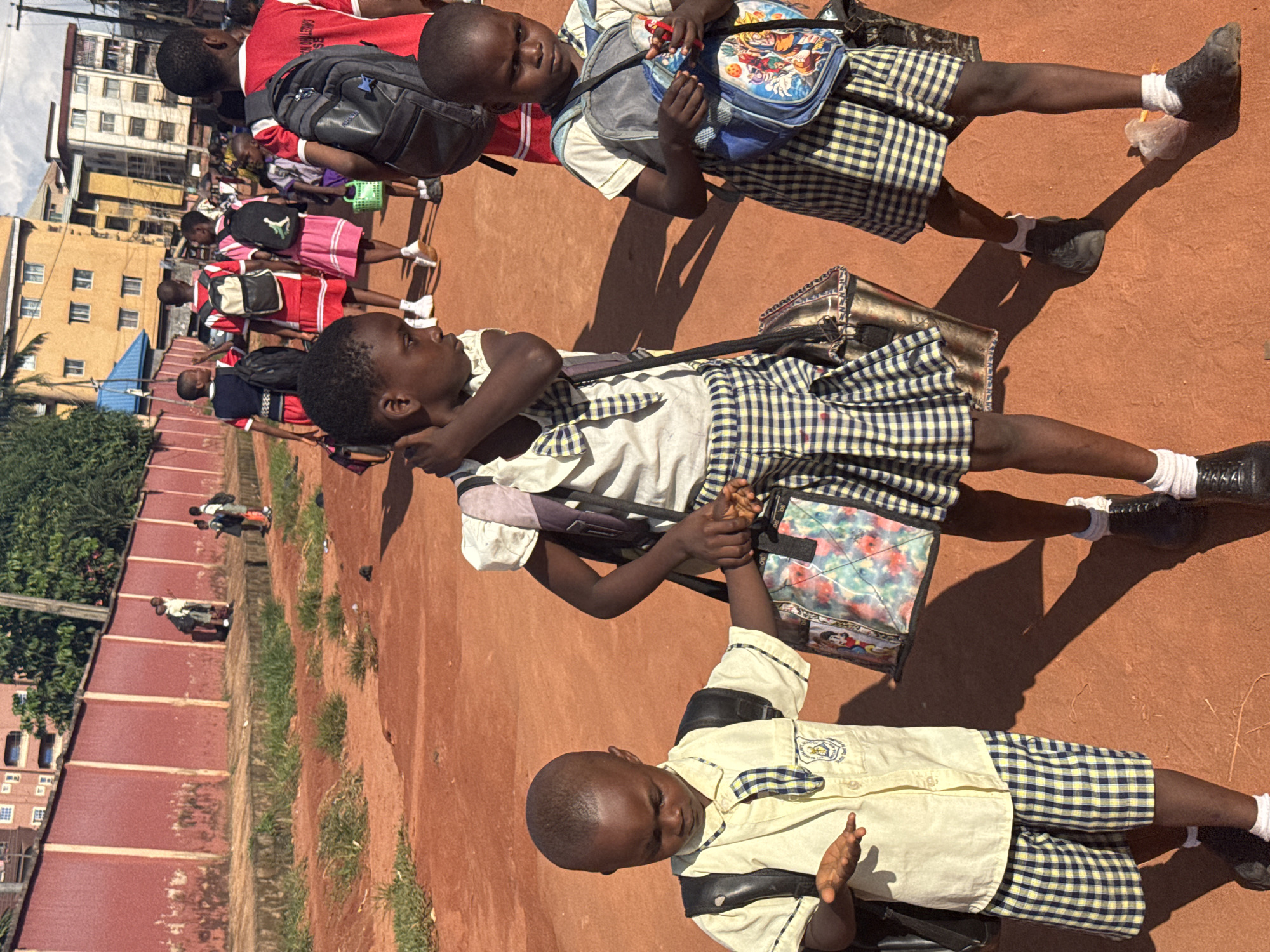 The image shows a group of children in school uniforms walking on a dirt path. The children are wearing plaid skirts and shirts, and some are carrying backpacks. In the background, there are buildings and trees. The sun is shining, casting shadows on the ground. The overall impression is of a typical school day in Onitsha, Nigeria.
The subjects are primarily children, likely students, dressed in distinct school uniforms consisting of plaid skirts and light-colored shirts. They are carrying various types of bags, including backpacks and satchels, some of which are decorated with cartoon characters. The scene is an outdoor setting, characterized by a reddish-brown dirt ground that appears to be a path or field. In the background, buildings are visible, suggesting an urban or semi-urban environment. Some greenery, including trees and grass, is also present, bordering the path.
The activity depicted is children walking, likely to or from school, or participating in a school-related event. Their posture and the direction of their movement suggest they are in procession or moving as a group.
Notable details include the time of day, indicated by the strong shadows cast by the children and the bright lighting, suggesting it is daytime, possibly mid-morning or afternoon. The weather appears to be sunny and warm. The location cues, such as the buildings and the dirt path, are consistent with an environment found in Onitsha, Nigeria. The children's expressions are not clearly discernible, but their body language suggests they are engaged in the activity.
There is no visible text in the image that is clear enough to be transcribed.