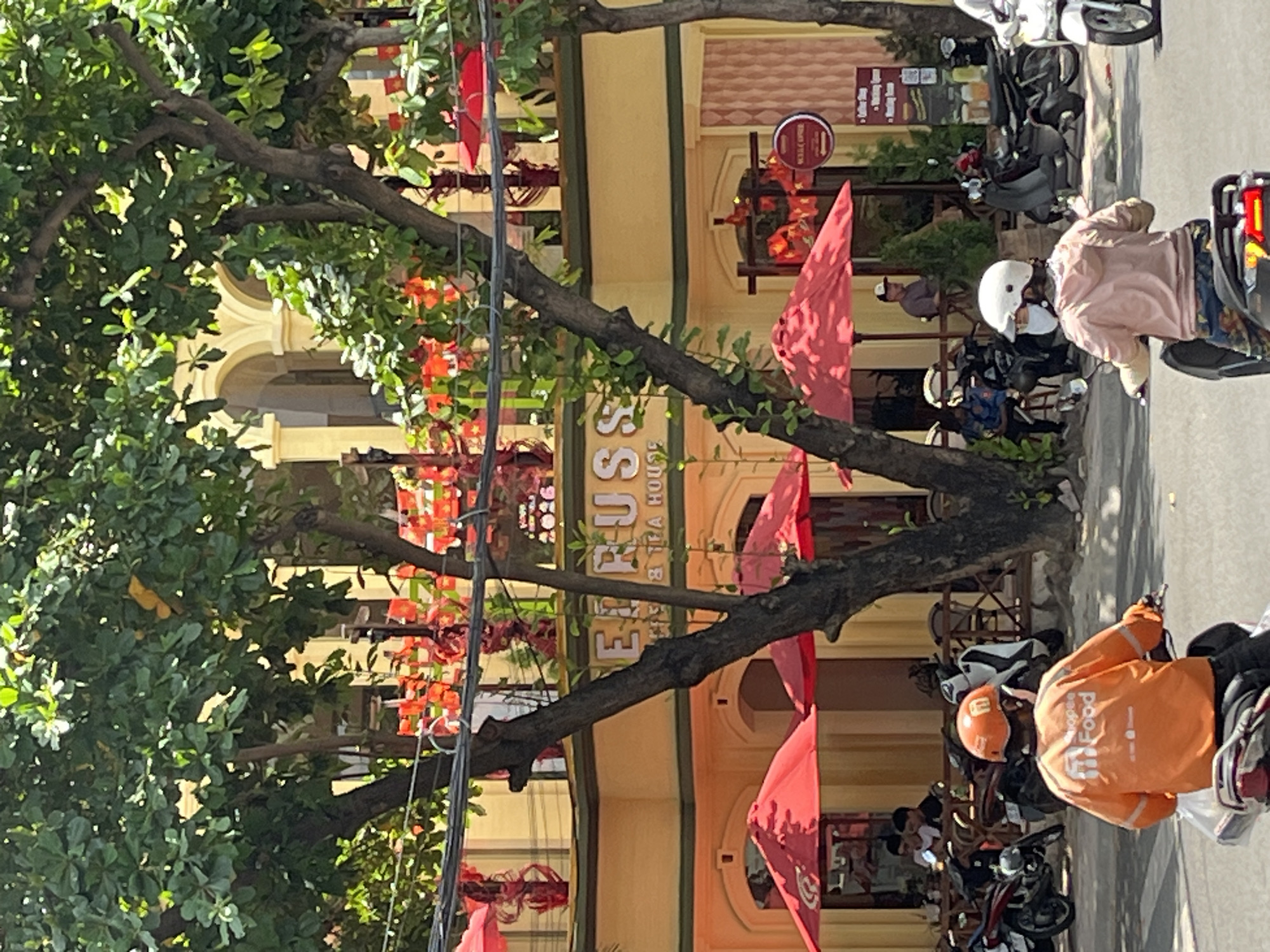 In a street scene in Nha Trang Ward, Vietnam, a Shopee Food delivery rider wearing an orange helmet and jacket is riding a motorcycle. Behind him, another person on a motorcycle wears a white helmet and a light pink hoodie. The background features a yellow building with arched entrances and green trees. Red umbrellas are set up outside the building, suggesting it is a cafe or restaurant. Several other motorcycles are parked. Red decorations are visible strung across the upper part of the building. The day appears sunny, with shadows cast on the street.