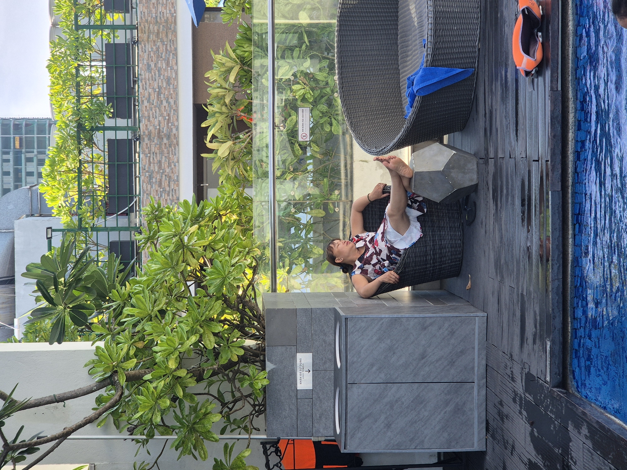 The image depicts an outdoor seating area adjacent to a swimming pool, likely within a hotel or resort in Đà Nẵng, Vietnam. A woman, appearing to be of Vietnamese ethnicity, is seated in a wicker chair with her legs extended, resting on a geometric concrete object. She is wearing a floral print dress and appears to be relaxed. To her left is a grey stone cabinet with two doors and two circular openings on top, suggesting it may be a trash or towel disposal unit. Above the cabinet, a sign with Vietnamese and English text indicates "USED TOWELS" with a downward arrow. Behind the woman, a clear glass railing separates the seating area from lush greenery and the upper levels of a building. In the background, several potted plants are visible on a balcony. A large, round, dark wicker sofa or lounger is to the woman's right, draped with a blue towel. The wooden decking around the pool area appears wet. An orange life vest lies on the deck near the edge of the pool. The overall atmosphere suggests a leisure setting in a tropical urban environment.
