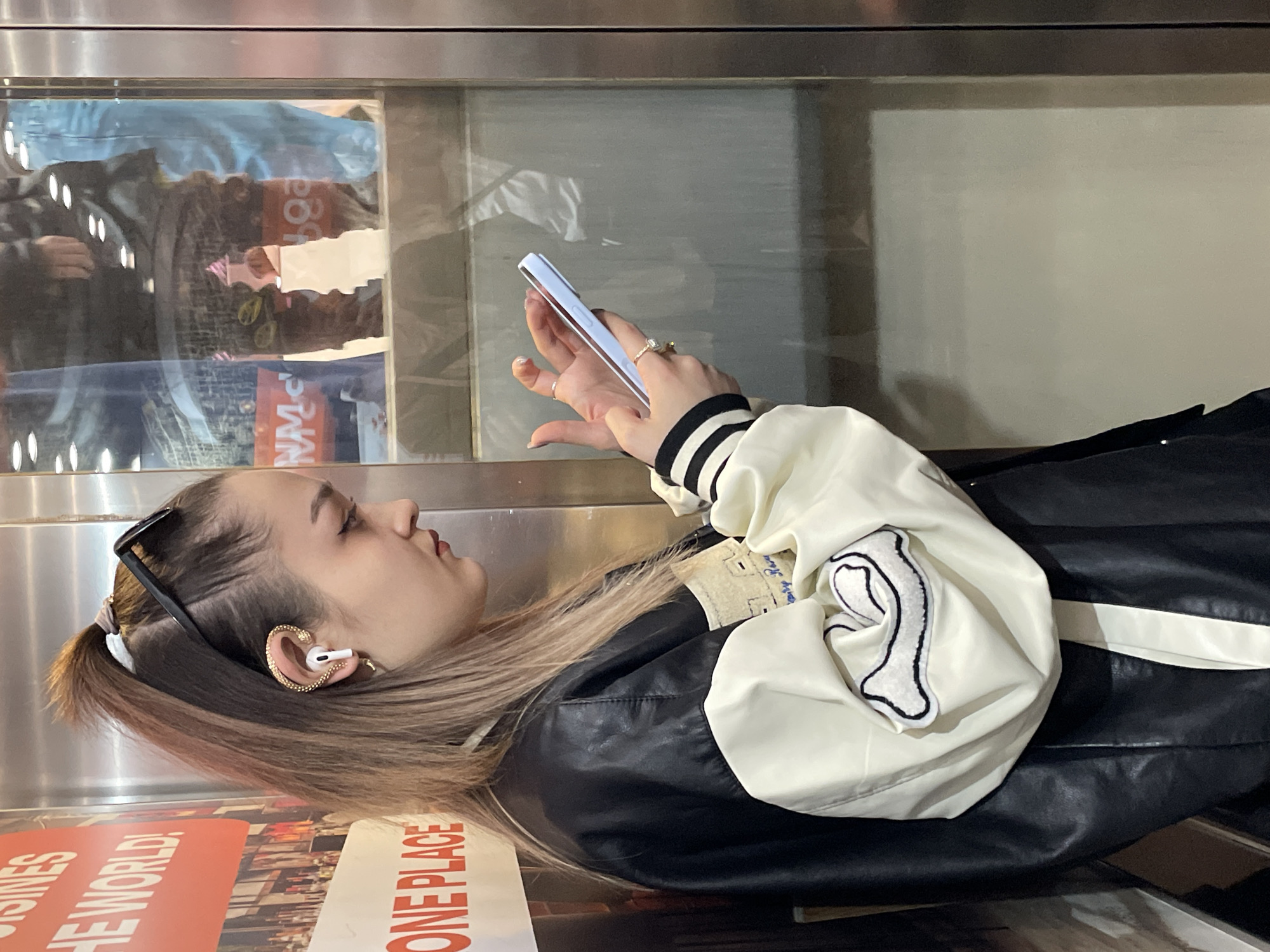 A young woman in a black and white letterman jacket is looking at her phone in an elevator. She has sunglasses in her hair, and an earbud in her ear. The elevator walls are stainless steel, and there is an advertisement for "One Place" and "Businesses of the World" on the wall. The reflection in the elevator shows people and merchandise, likely in a store. There are no specific details that would definitively place this image in Saint Petersburg, Russia.