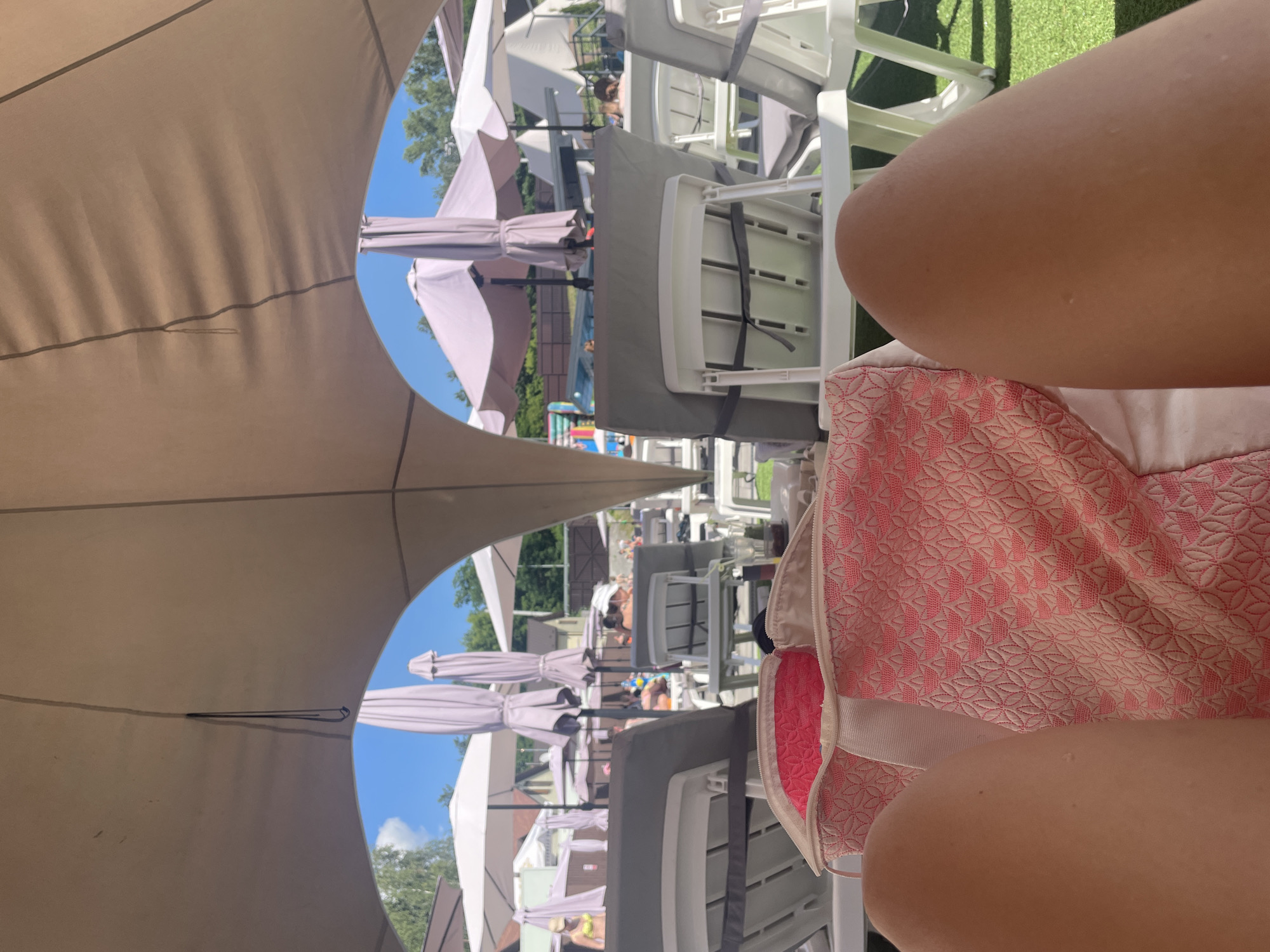 The image depicts a first-person perspective at a sunny outdoor swimming pool area in Zazymia, Ukraine. The foreground is dominated by a pink and white patterned beach bag and the bare legs of the photographer. In the midground, rows of empty, grey-cushioned lounge chairs are set up on a green artificial turf surface. Behind the chairs, a variety of light-colored umbrellas provide shade, with several closed and a few open. Beyond the umbrellas, there are trees and buildings, suggesting a resort or recreational facility. The sky is clear and blue with some scattered clouds. Several people can be seen in the background, some relaxing on chairs, others near the pool or in a shallow water area. The overall impression is one of a leisurely summer day at a poolside resort.