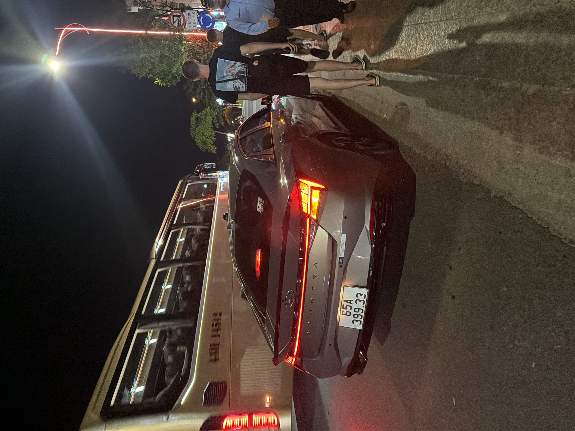 This nighttime image depicts an urban street scene in Nha Trang Ward, Vietnam. A grey Hyundai Elantra sedan, identified by its illuminated "ELANTRA" badging and distinctive full-width LED taillight bar, is positioned in the mid-ground. Its Vietnamese license plate, "65A 399.33," indicates registration in Can Tho province. To the left, a large, light-colored passenger bus with the numbers "43H 145.12" visible on its side is partially obscured and appears to be in motion. On the right, a bright streetlight with yellow-white illumination emanates from a red-painted pole, casting long shadows of pedestrians onto the sidewalk. Below the streetlight, multiple street signs are visible: a blue circular sign with a white arrow pointing right, and a white rectangular sign with red text displaying time intervals "6:30 - 7:30" and "14:30 - 18:30," likely indicating traffic restrictions. Three adult males are walking on the sidewalk towards the right side of the frame. The closest man is seen from behind, wearing a black t-shirt with a graphic print, black shorts, and sandals, holding a smartphone. Another man in dark clothing is partially visible behind him. Furthest right, a man in a light blue collared shirt and dark trousers, resembling a uniform, is also walking. The ground consists of asphalt for the road and a paved sidewalk.