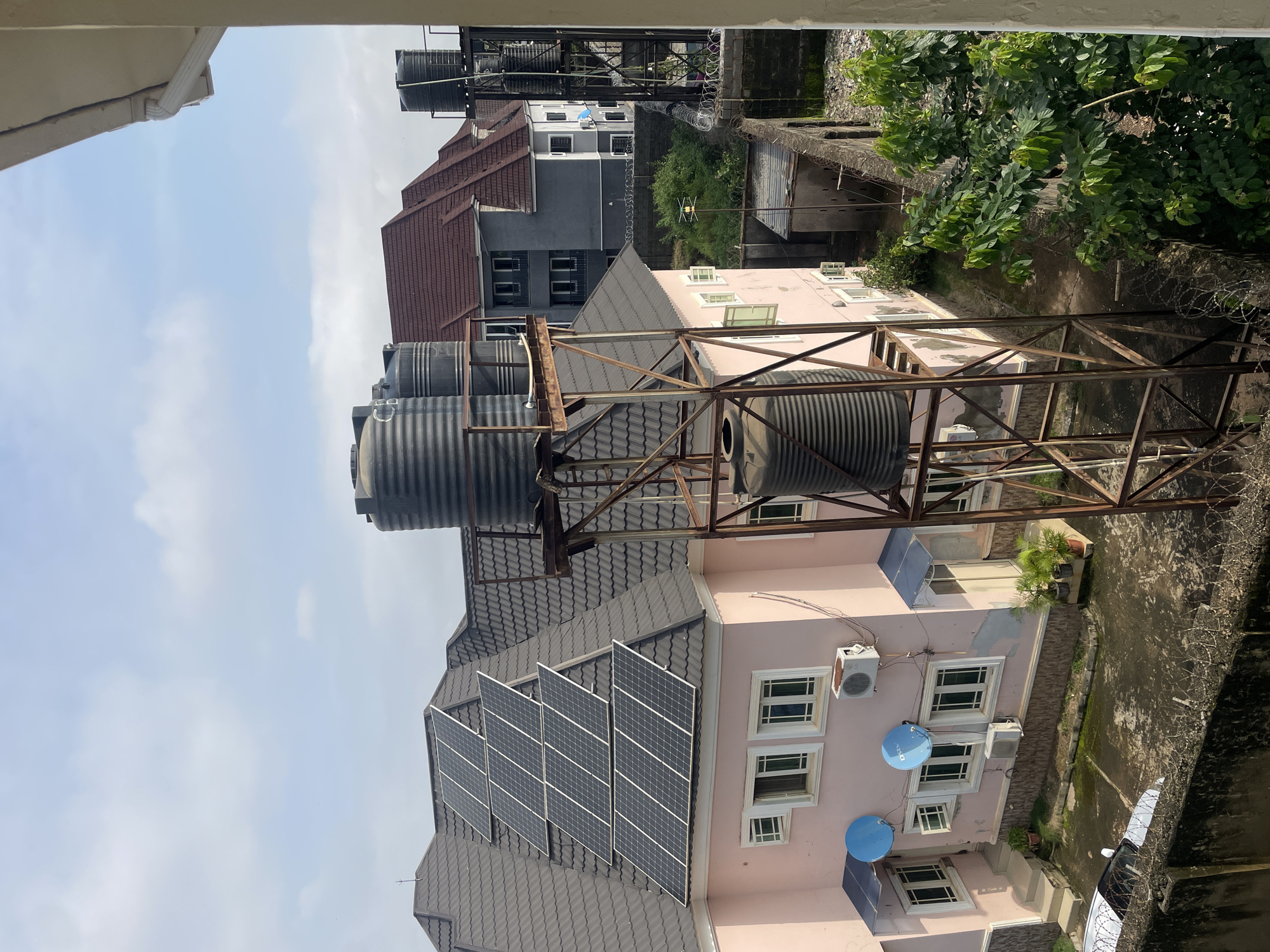An aerial view captures a residential area in Abuja, Nigeria. The foreground features a light pink, two-story building with a dark gray, tiled roof adorned with solar panels. Several large, black water tanks are elevated on a rusty metal structure in front of the building. Two blue satellite dishes are mounted on the building's exterior, along with air conditioning units. The building has multiple windows and doors, and a white car is parked in the paved courtyard below. A concrete wall topped with barbed wire separates this property from neighboring areas. In the background, other residential buildings with different roof colors and architectural styles are visible, along with lush green vegetation and trees. The sky is partly cloudy.