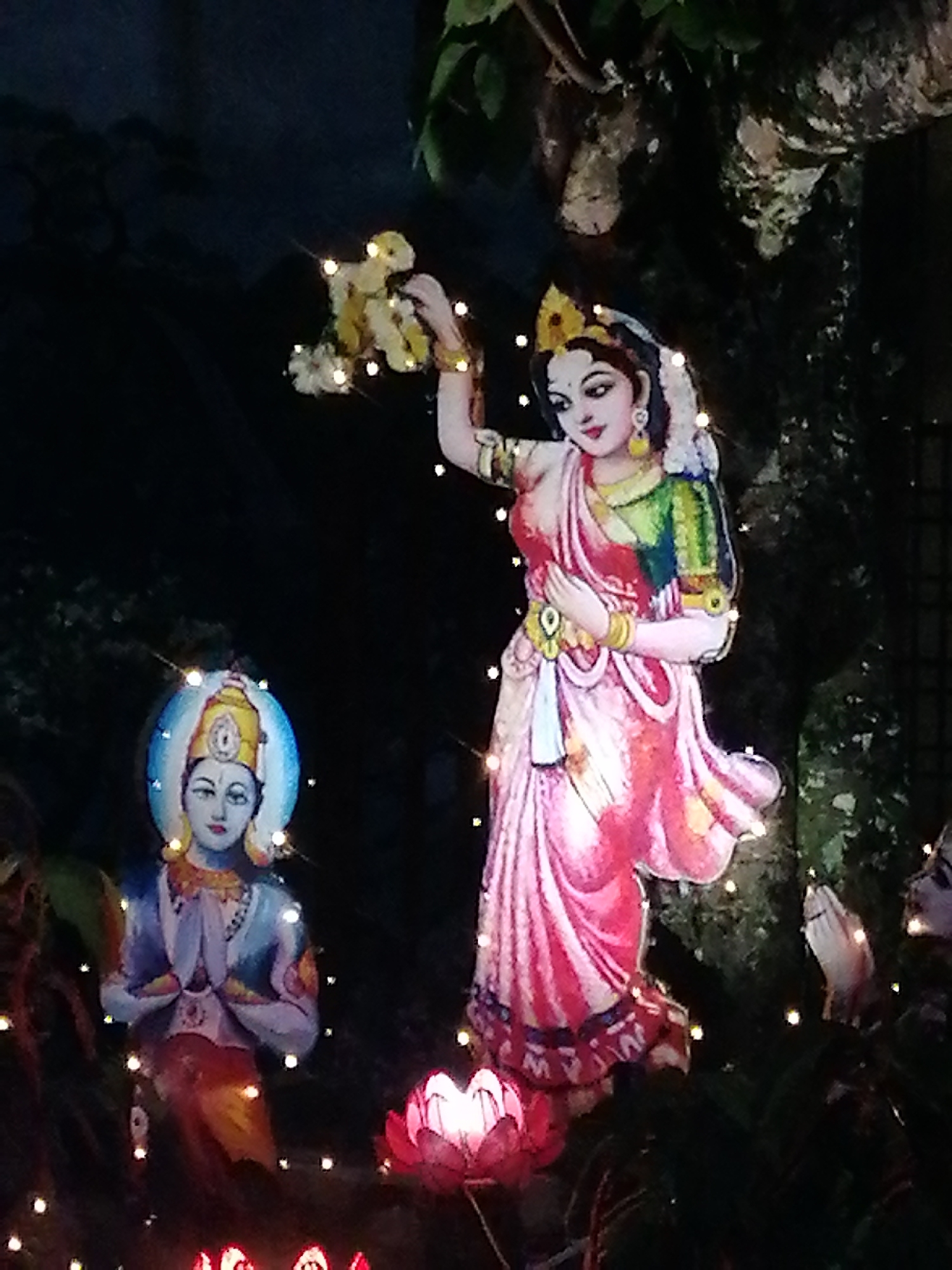 The image captures a nighttime scene featuring religious imagery, likely in Da Lat, Vietnam. At the forefront are two illuminated cardboard cutouts: one of a female figure in a red and pink sari holding a bouquet of flowers, and another of a male figure with a blue halo around his head in a prayerful pose. The cutouts are surrounded by a series of small, warm-toned lights, some arranged within lotus flower-shaped light fixtures. Behind these figures, there appears to be a dark, forested or garden environment, possibly a tree. The overall atmosphere is festive and devotional.
