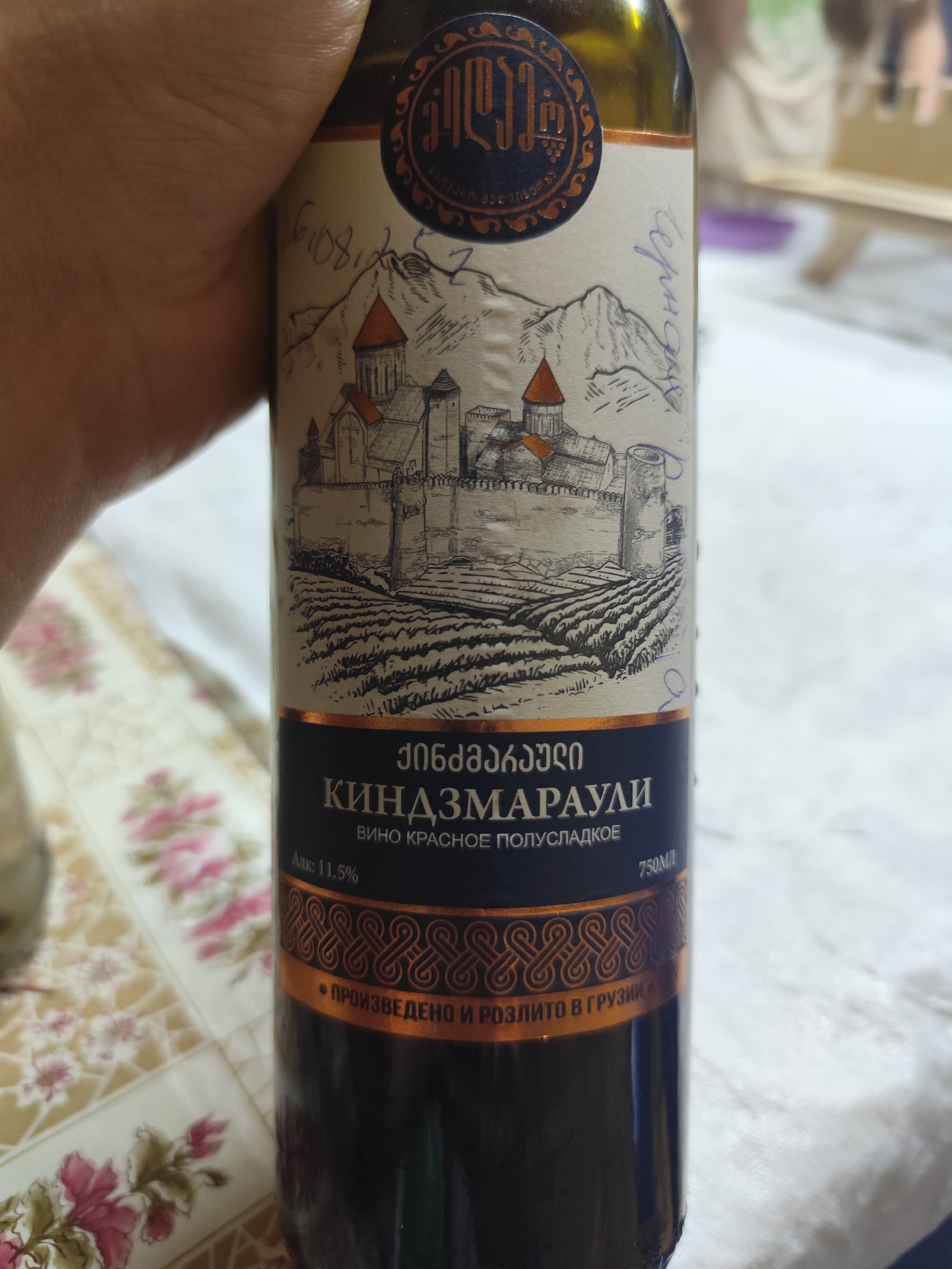 The image shows a bottle of wine with a label that is partially obscured by a hand. The label features an illustration of a castle and mountains, along with Georgian and Russian text. The text on the label identifies the wine as "Kindzmarauli" and indicates it is a semi-sweet red wine with an alcohol content of 11.5%. The bottle is on a table covered with a floral patterned tablecloth. The location in Kolpakovo, Russia is not visible in the image.