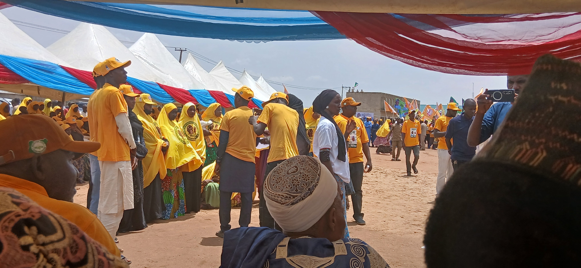 In Rigachikun, Nigeria, a political gathering is taking place outdoors under a canopy of red, white, and blue fabric. A crowd of people, predominantly dressed in yellow and orange attire, are milling about. Many wear yellow headscarves, and some have yellow caps. Several individuals are holding flags and banners, indicative of a political rally or event. In the foreground, partially obscuring the view, is a person wearing a patterned headdress and a dark blue garment. Another person in the middle ground is actively taking a photo with a smartphone. Tents are visible in the background, suggesting a temporary setup for the event. The ground appears to be sandy or dusty.
