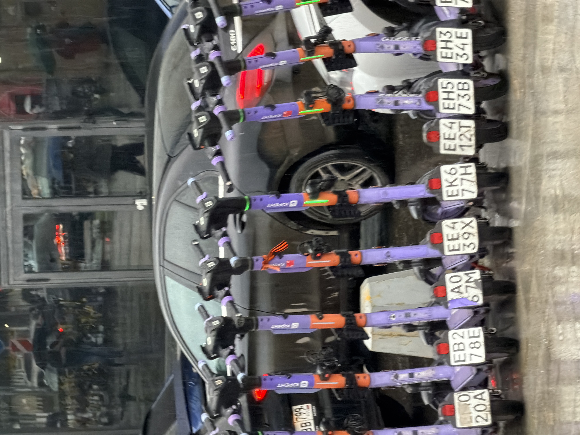 The image shows a row of electric scooters parked next to a car, presumably in a city setting. The scooters are purple and orange with black handles, each sporting a license plate. The plates show codes, such as "20A," "78E," "67M," and so on. The scooters are arranged closely together, indicating a designated parking area. The car is a dark color, possibly black or dark gray, and appears to be parked on the street alongside the scooters. In the background, there is a building with glass windows, reflecting the urban environment. The presence of license plates suggests a regulated scooter-sharing system. Given the OCR text with "KOPIEHIT," this is likely a scene in Moscow, Russia.