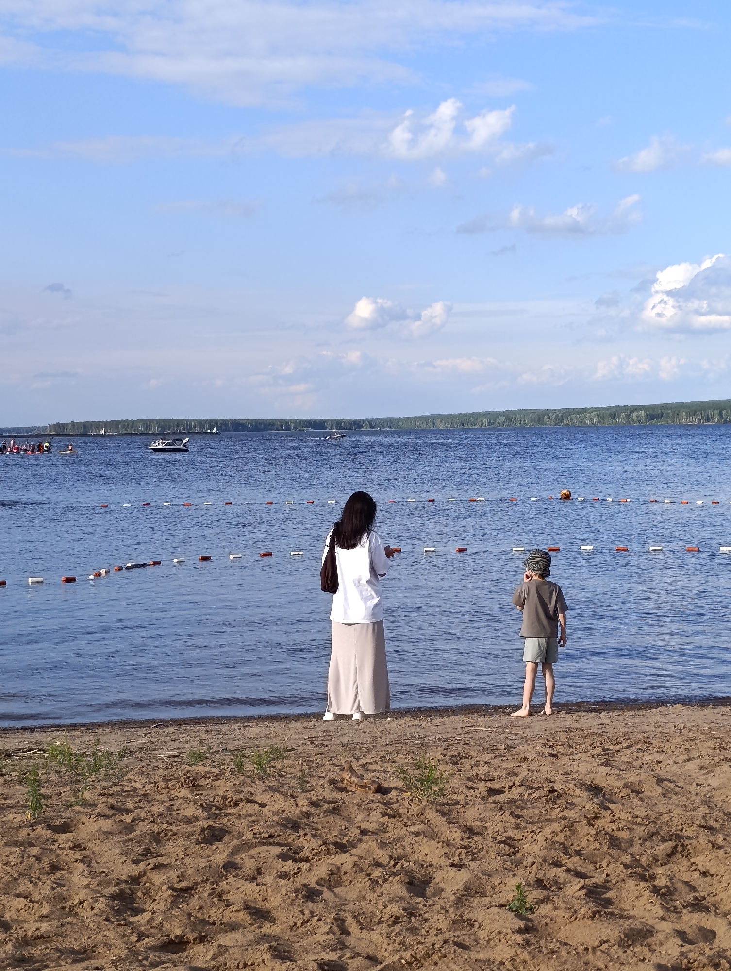 A wide shot captures a serene summer day at a beach in Chaikovsky, Russia, bordering the Kama River. Two figures, a woman and a child, stand near the water's edge, facing away from the camera, contemplating the vast expanse of blue. The woman, dressed in a white shirt and a long, flowing skirt, holds a dark bag over her shoulder. The child, in a grey t-shirt and shorts, wears a patterned hat.
In the background, the calm waters of the Kama River stretch towards a distant, tree-lined horizon. Several boats, including sailboats and a motorboat, dot the surface of the river, suggesting recreational activities. A floating rope with buoys marks a boundary in the water. The sandy beach in the foreground is dotted with sparse green vegetation. The sky above is a brilliant blue, adorned with scattered white clouds, casting a gentle light on the scene. The overall atmosphere is peaceful and tranquil, indicative of a perfect summer's day by the river.