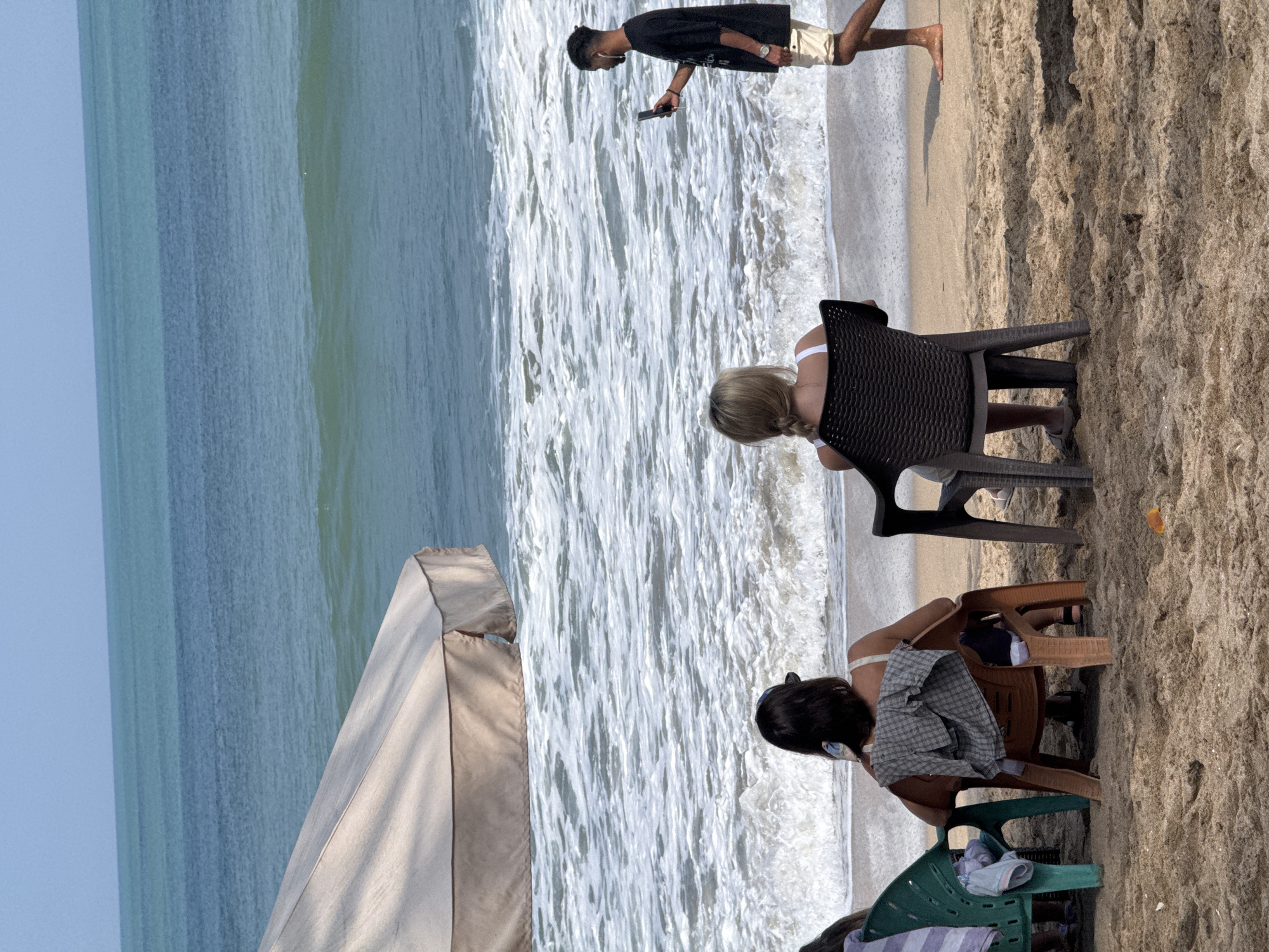 In Kuta, Indonesia, two women sit in chairs on a sandy beach facing the ocean. A large beige umbrella is partially visible to the left. The ocean has white waves and a clear blue sky above. A man walks along the shore in the background, holding a phone to take a picture. The woman on the right has blonde hair and wears a white top. The woman on the left has dark hair and a gray checkered shirt draped over her chair. Another person is partially visible in a green chair to the far left. The sand is textured with shadows.