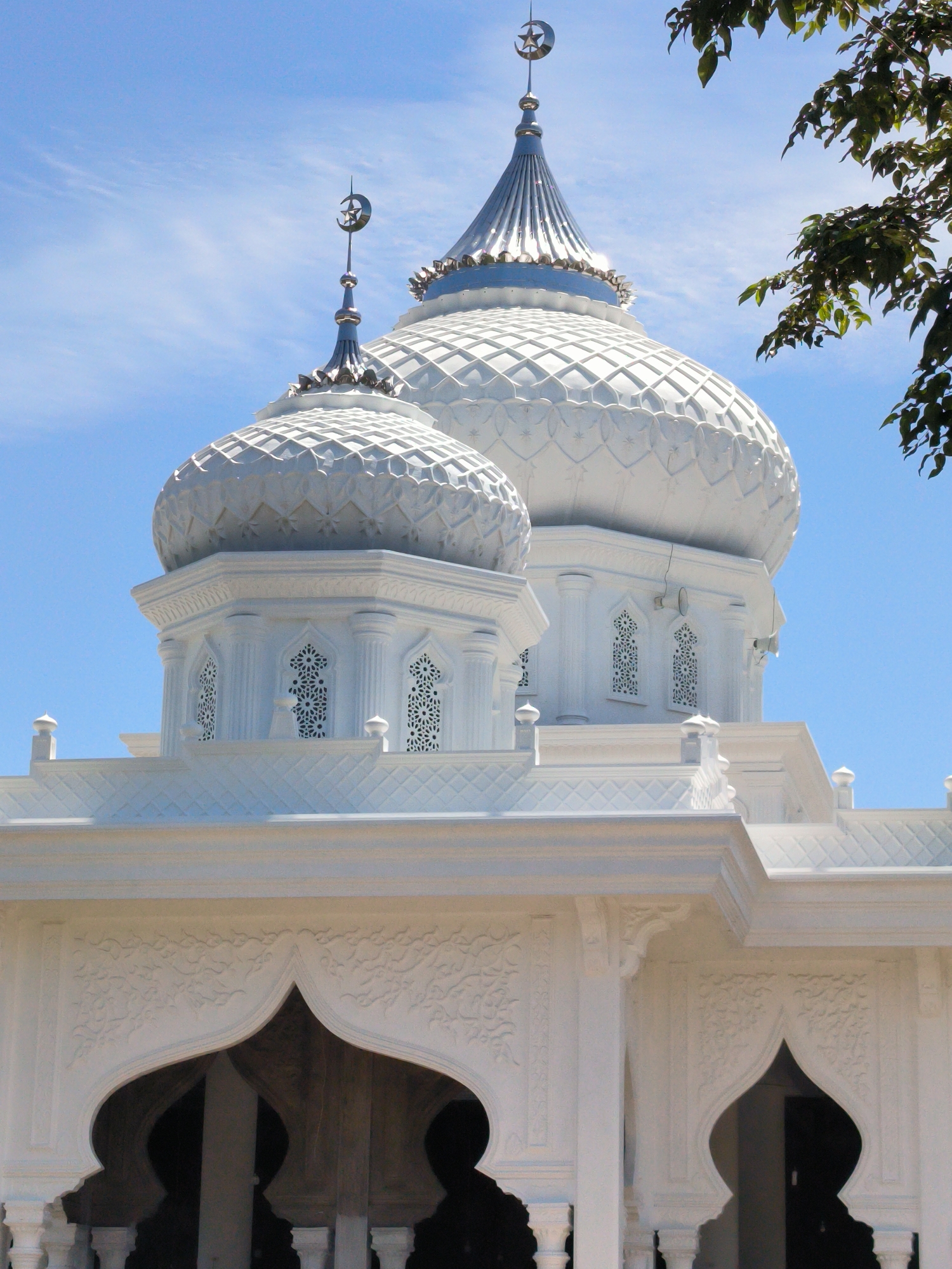 The image shows a close-up of the Baitul Makmur Mosque located in Meunasahgeumata, Indonesia. The mosque is predominantly white, with two domes visible. The larger dome has a pointed silver top, each topped with a crescent moon and star. Decorative patterns are visible on the domes, and the architecture includes arches and ornate carvings. A blue sky is in the background. A tree branch with green leaves is visible in the upper right corner of the image.
