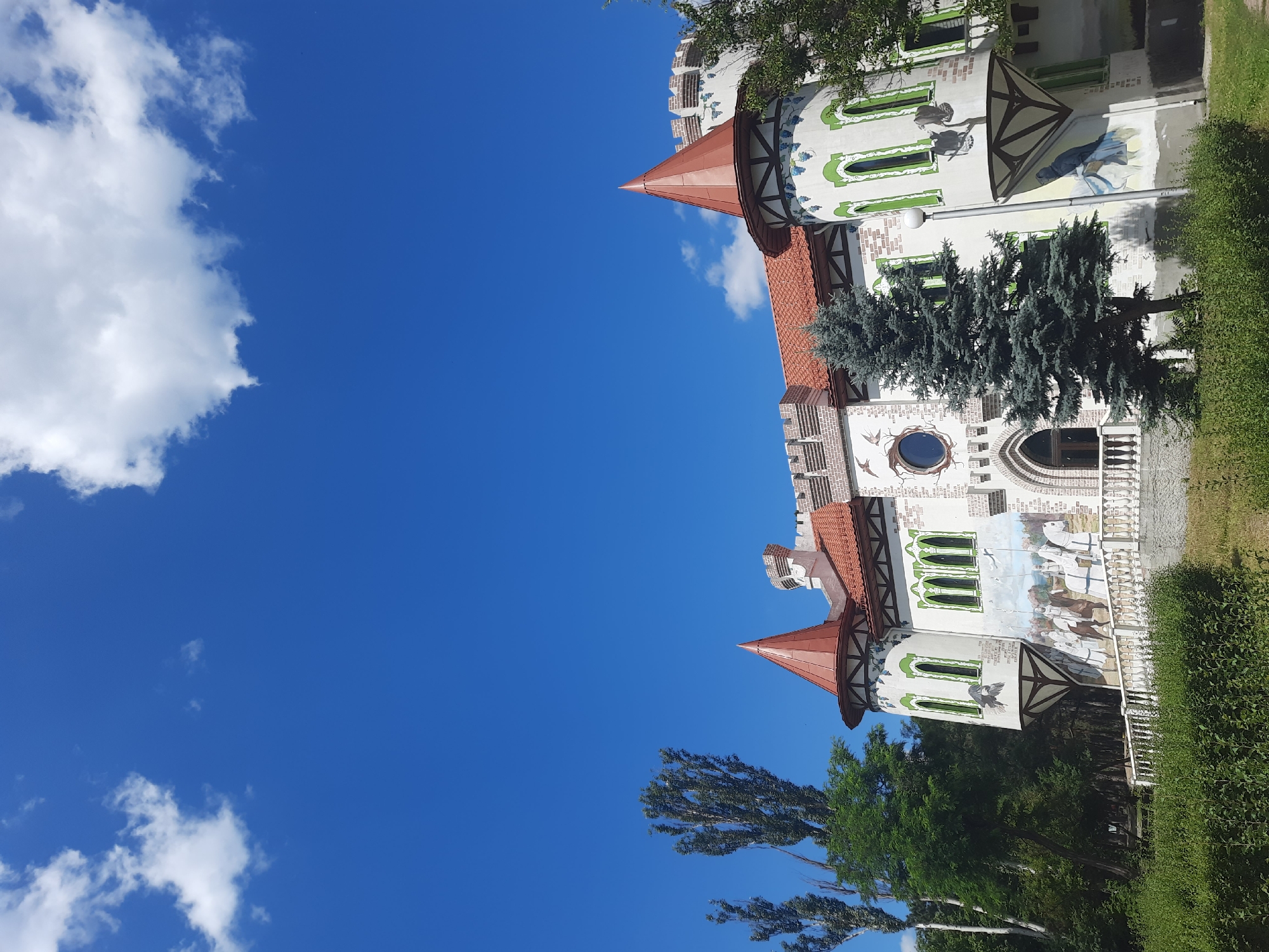The image shows a fairy-tale-like castle with red conical roofs, white walls decorated with murals, and green-trimmed arched windows. The castle is surrounded by lush green trees and bushes, with a white railing in the foreground. The sky above is a vibrant blue with scattered white clouds. The castle appears to be a tourist attraction or a whimsical building, possibly a part of a park or entertainment venue. This castle is known as the "Fairy Tale Castle" and is located in the city of Horishni Plavni, Ukraine. The murals on the castle depict scenes of knights on horseback and various birds, enhancing its fantastical theme.