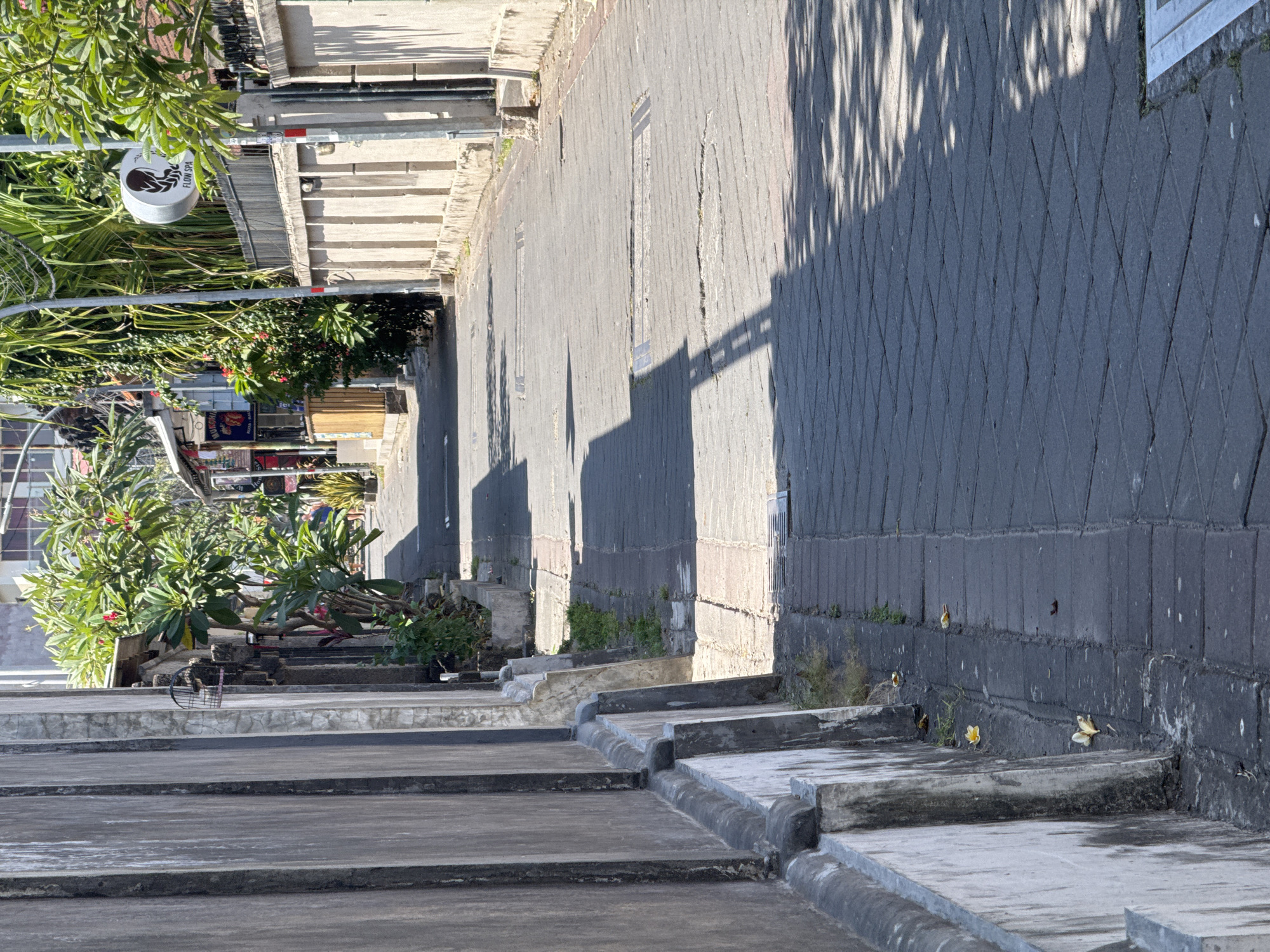 The image shows a narrow street in Kuta, Indonesia, lined with buildings and vegetation. The street is paved with dark gray interlocking stones, with a lighter colored brick path running along the left side. A tall, textured concrete wall runs along the left edge of the street, with several vertical concrete posts extending upwards. Greenery, including trees and bushes with red flowers, grows along the buildings on the right side of the street. A sign for "FLOW SPA" is visible on the right, attached to a fence. Shadows from trees and buildings cast across the street. A few fallen yellow flowers are scattered on the pavement. The scene appears to be during the daytime, with bright sunlight.
