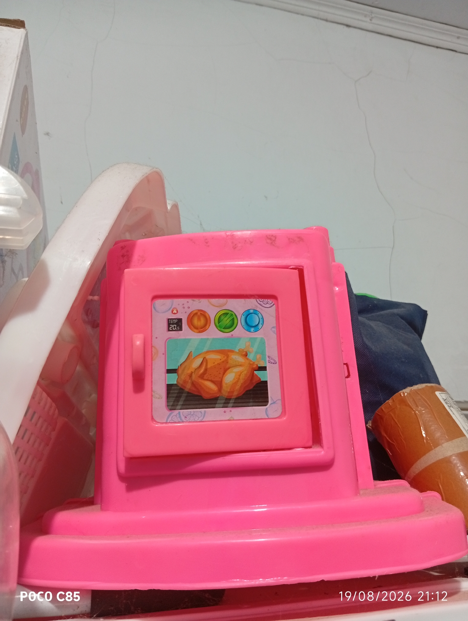 A low-angle, eye-level shot captures a pink toy oven with a digital display showing "TEMP 20S" and a rotisserie chicken graphic inside. The oven is on a shelf against a white wall with visible cracks. To the left of the oven is a white, curved plastic object, possibly part of another toy. To the right, a dark blue fabric item and a brown cylindrical object, likely a roll of tape or paper, are partially visible. The bottom of the image shows the base of the toy oven and a watermark reading "POCO C85" and "19/08/2026 21:12". The scene appears to be an indoor domestic setting in South Tangerang, Indonesia.