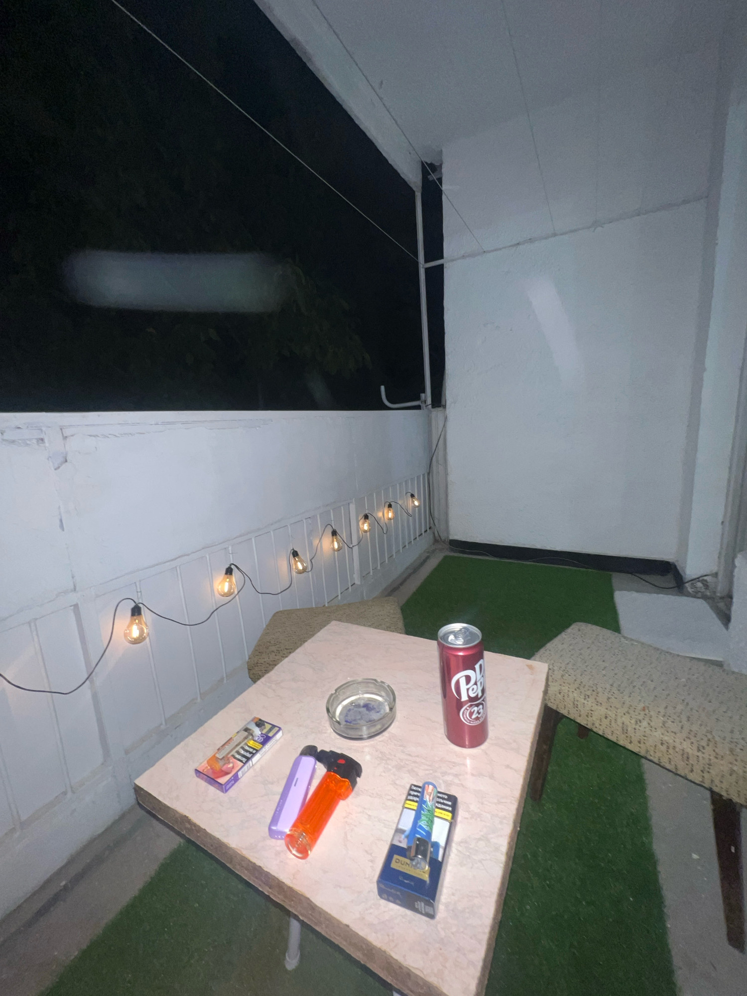 The image depicts a dimly lit balcony at night, likely in Montana, Bulgaria, based on the available context. The balcony is furnished with a light-colored marble-patterned table, an ashtray, a can of Dr Pepper, two lighters (one orange, one purple), and a pack of cigarettes on the table. To the right, a cushioned bench is partially visible. A string of vintage-style light bulbs hangs along the white railing, casting a warm glow. The floor is covered with artificial grass. The background shows dark foliage and a white wall. There are no people visible in the image, and no specific actions or interactions are evident.