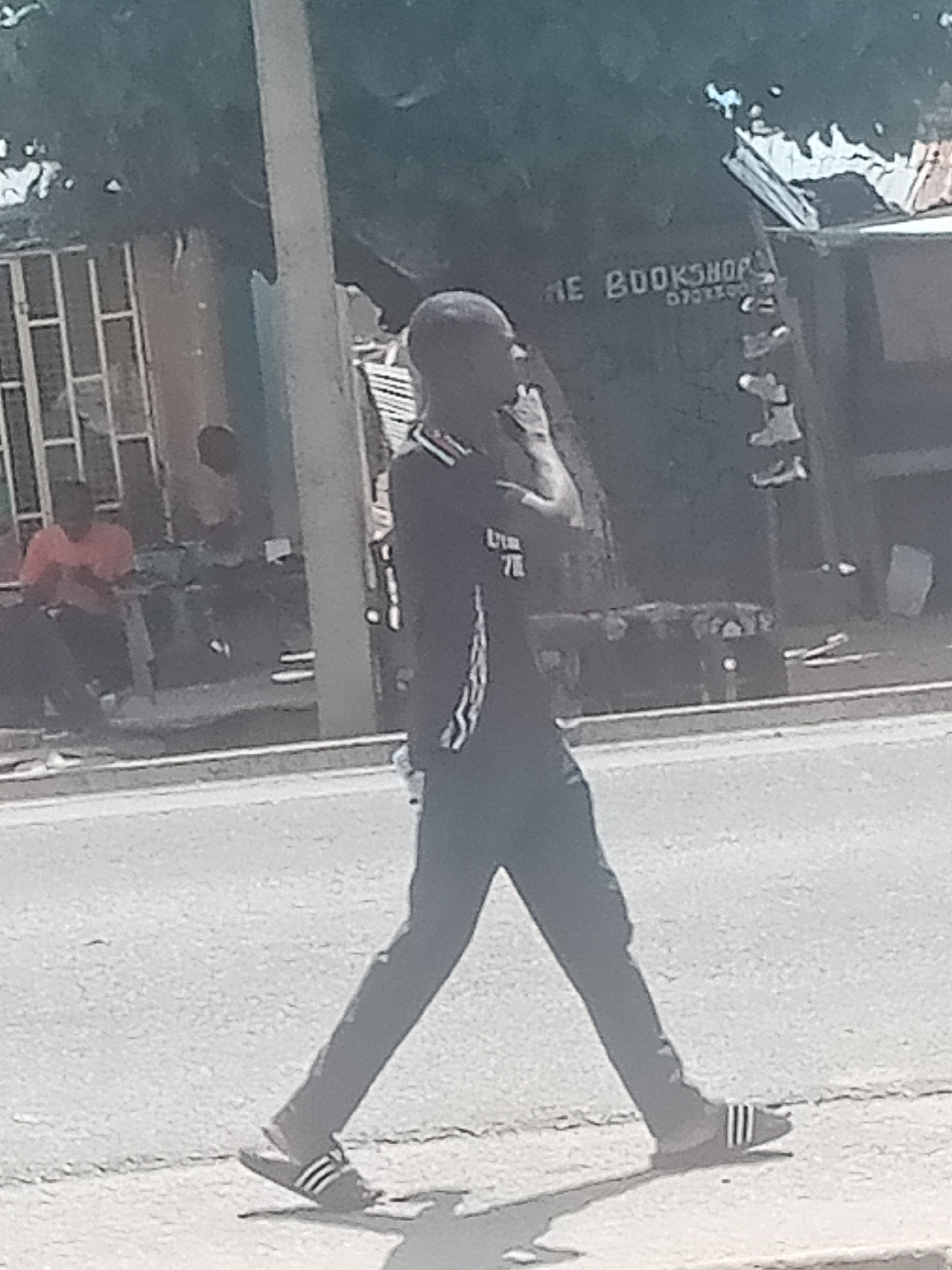 In Jalingo, Nigeria, a lone individual walks across a paved street. The person, clad in a dark t-shirt with a white stripe down the sleeve and dark pants, is wearing Adidas slide sandals. Behind the walker, a narrow sidewalk runs parallel to the street, bordered by a grassy verge. Further back, a light-colored wall with a barred window is visible, and to the right, a building with signage that reads "THE BOOKSHOP" and a phone number is present. The scene is bathed in daylight, casting shadows on the ground. Other indistinct figures and structures are visible in the background, suggesting an urban environment.