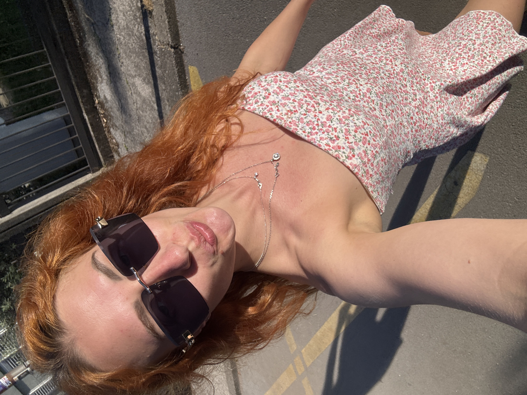 A young adult female with long, reddish-auburn hair is depicted in a selfie taken outdoors in the city of Budva, Montenegro. She is holding the camera at arm's length, angled slightly downwards, and is puckering her lips in a pose. Her face is partially obscured by rectangular, dark-lensed, rimless sunglasses with gold-tone accents on the upper corners and hinges, one of which has "Cartier" branding visible. She wears two silver-tone necklaces: an upper, shorter chain with small chevron-like elements, and a lower, longer chain featuring a circular, faceted pendant. Her skin appears fair, with some sun-kissed redness on her forehead and cheeks. A small, light brown mole is visible on her inner right arm. She is wearing a white, strapless or thin-strapped short dress adorned with a dense pattern of small pink and green floral elements. The background shows a dark gray paved surface, likely asphalt or concrete, exhibiting cracks and some scattered debris. Yellow painted lines are visible on the ground. To the upper left and right, a dark metal fence or railing is present, with green foliage visible behind it on the upper right. Strong sunlight casts distinct shadows, indicating a bright outdoor setting.