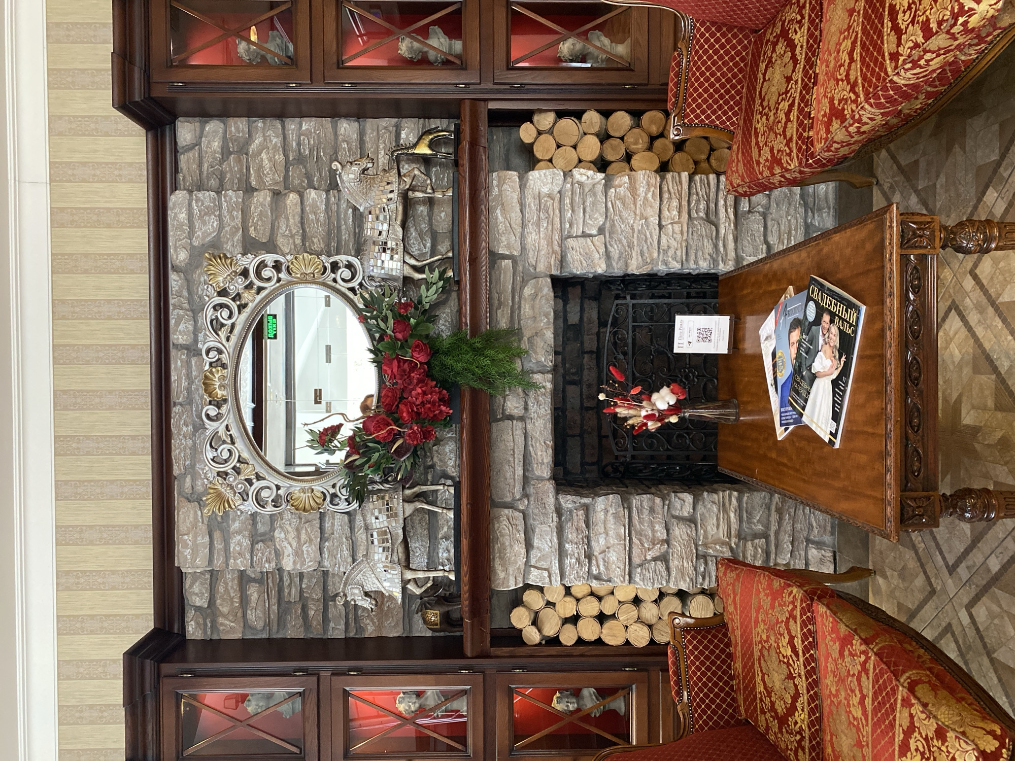 The image captures an interior scene, likely a decorative room or lounge. The focal point is a fireplace structure embedded in a wall unit, incorporating stonework and decorative elements. The fireplace itself has a black metal grate and is flanked by stacks of firewood on both sides. Above the fireplace, a decorative oval mirror with ornate gold and silver detailing is mounted on the stonework, adorned with floral arrangements in red and green.
The wall unit surrounding the fireplace includes built-in cabinets with glass-fronted sections displaying figurines. On a table to the right, several magazines are placed. Two ornately patterned, red and gold sofas are partially visible, adding to the overall richness of the interior. The wallpaper behind the structure has a striped pattern in neutral tones. The flooring visible near the sofas has a geometric, tiled pattern.
Based on the Cyrillic text on the magazines, which includes "Свадебный Вальс" (Wedding Waltz), and the description provided of the location being in Yekaterinburg, Russia, the image is likely taken in a space associated with wedding planning or events. The overall aesthetic of the room suggests a style that blends classical and rustic elements.