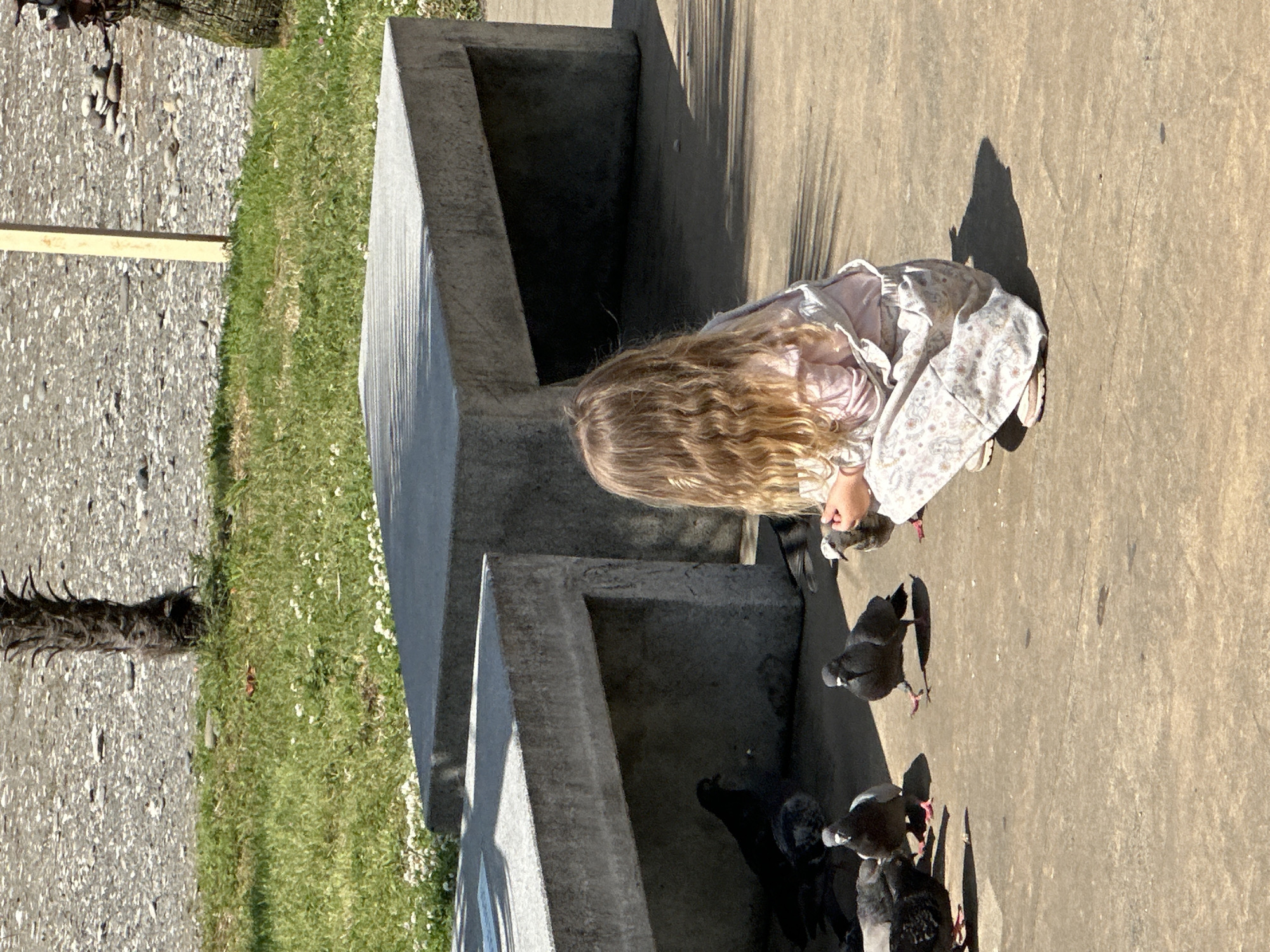 A girl in a light-colored dress and long blonde hair is crouched down on a paved surface, possibly a sidewalk or plaza. She is surrounded by several pigeons that are clustered around her. The background features a concrete structure with stepped levels and green grass visible to the left. The scene is outdoors and lit by sunlight, with shadows cast by the girl and the surrounding structures. The location is likely in a park or public space in Batumi, Georgia, given the style of the concrete structure.