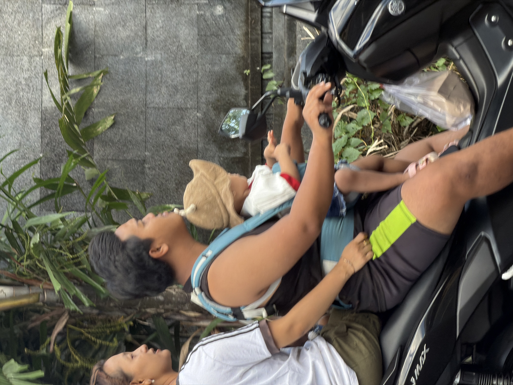 The image depicts an outdoor scene in North Kuta, Indonesia, featuring a family of four on a black Yamaha NMAX scooter. The photo appears to be taken during the daytime, with natural lighting.
In the foreground, an adult male, wearing a dark tank top, is seated on the scooter. He has a baby or young child in a blue and grey forward-facing carrier on his chest. The baby wears a white top and a light brown hat, possibly with animal ears, and is barefoot. The male's hands are on the scooter's handlebars. Behind him, an adult female is seated on the scooter, wearing a white t-shirt with dark stripes on the sleeves. Her hair is light brown or auburn. Further back on the scooter, a second child or person is visible, wearing dark shorts with a neon green stripe on the side.
The scooter itself is black, with "YAMAHA" visible near the handlebars and "NMAX" emblazoned on the side panel. A transparent plastic bag containing items rests on the scooter's footwell.
The background consists of a grey tiled or stone wall and lush green foliage, suggesting a tropical or subtropical environment. The overall activity appears to be a family outing or daily commute on the scooter, a common mode of transport in the region. All individuals seem relaxed.
