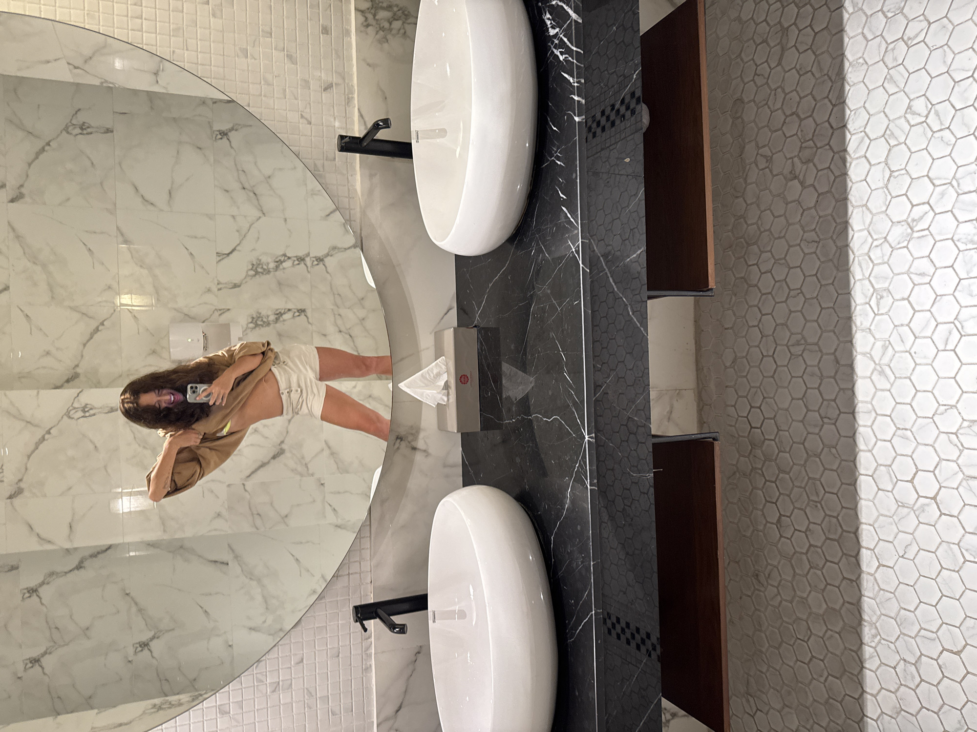 A vertical, medium shot depicts a bathroom interior with a large, round mirror reflecting a person taking a selfie. The person has long, wavy brown hair, and is wearing a tan shirt and white shorts. They hold a phone with their right hand, making a face towards the camera.
Two white, oval sinks are positioned below the mirror. Black faucets are installed on each sink. Between the sinks, a gray tissue dispenser is mounted on a black marble countertop. A dark wood cabinet stands beside the countertop.
The bathroom walls are tiled with white marble near the mirror and white hexagonal tiles on the right side of the frame. The tiles reflect light, creating a bright environment. The scene's location cannot be determined.