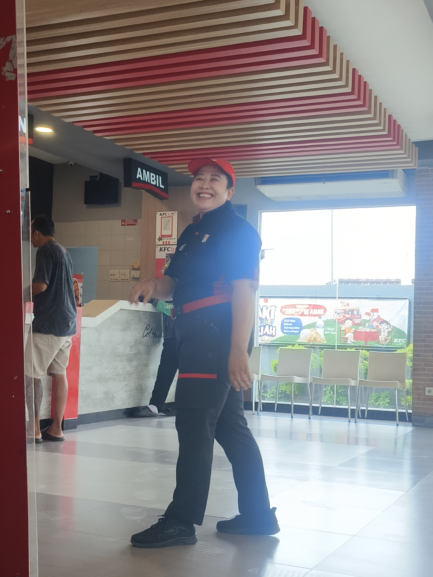 A woman in a red cap and KFC uniform, with a wide smile, stands indoors at a KFC restaurant. She is facing the camera, her arms outstretched and slightly blurred from movement, suggesting she might be gesturing or in motion. She is wearing black pants with red trim and black athletic shoes. Behind her, a man in a dark t-shirt and light shorts is facing away from the camera, looking at a counter. The environment appears to be a dining area or service counter, with a ceiling decorated with alternating red and wood-colored slats. A large window in the background shows a bright outdoor scene with white chairs lined up in front of it. A KFC banner with Indonesian text and cartoon illustrations of children is visible through the window. The floor is tiled in a pattern of light and dark gray squares. The overall impression is of a casual, well-lit restaurant setting.