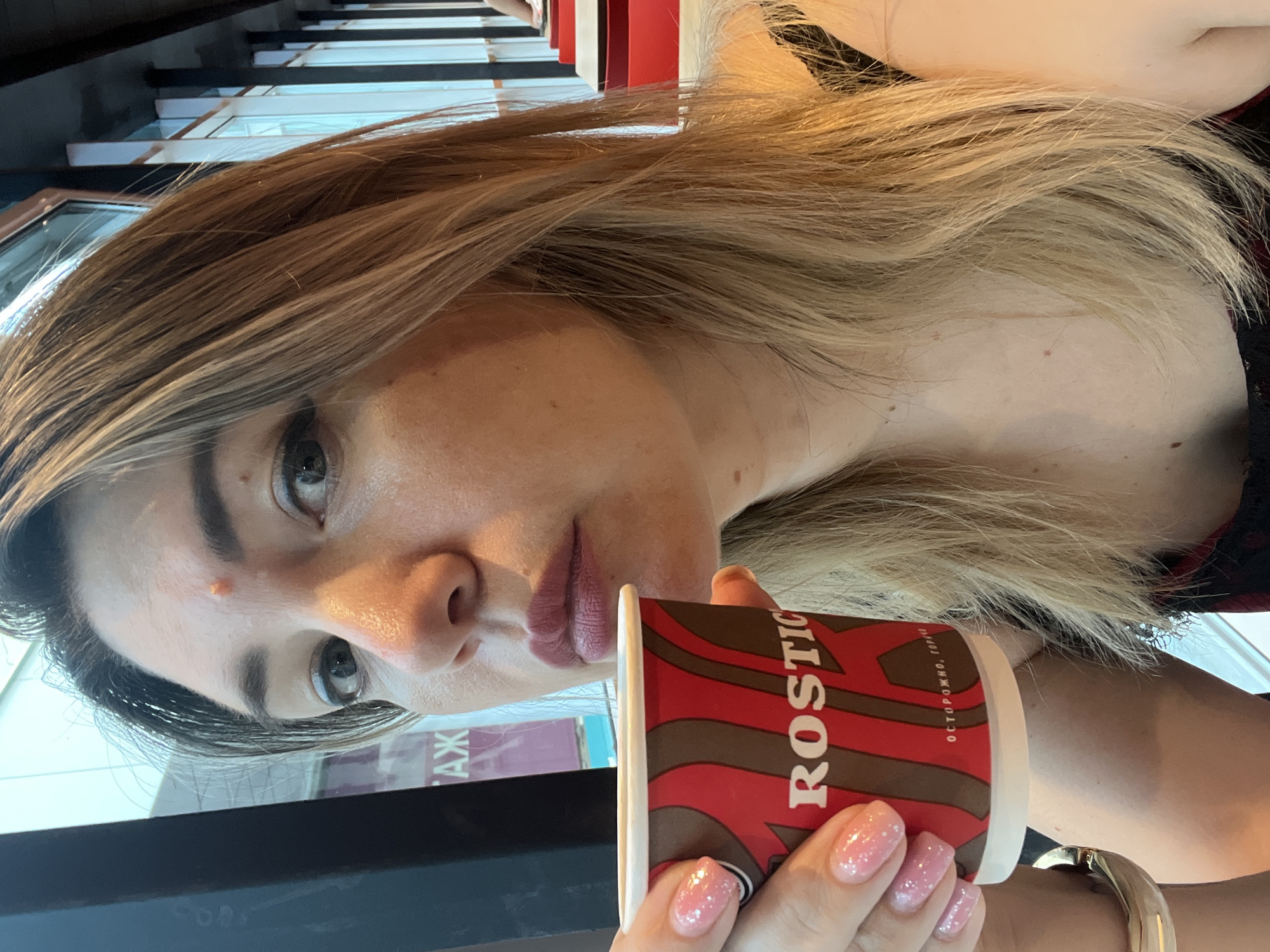 The image is a close-up, rotated selfie of a young woman holding a coffee cup. She has long, blonde, layered hair with streaks of lighter blonde. Her face is turned towards the camera, and she is looking directly into the lens with a neutral expression. She is wearing light makeup, including lipstick in a muted pink shade and subtle eye makeup. She has a small pimple on her forehead. Her nails are painted a shimmery pink.
She is holding a red and brown disposable coffee cup that has text on it. The visible text reads "ROSTIC" and in smaller Cyrillic letters below it, "ОСТОРОЖНО, ГОРЯ". This translates to "CAUTION, HOT". The background is blurred but appears to be an indoor setting with shelves containing items. The lighting suggests it could be daytime. The presence of the coffee cup and the casual pose imply the woman is likely at a cafe or a similar establishment. The context provided indicates the location is Sochi, Russia.