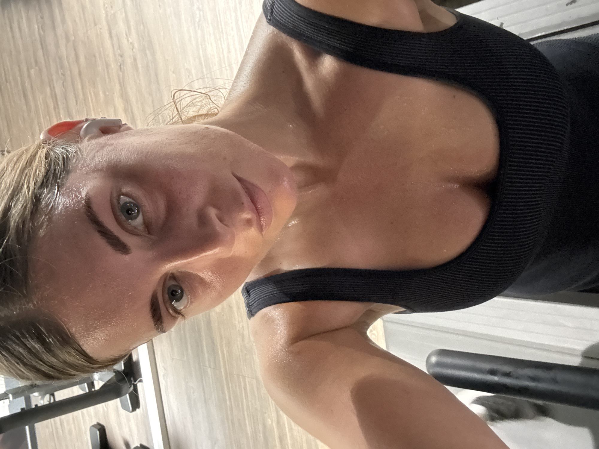 A close-up, tilted selfie captures a woman in athletic attire, presumably after a workout. She wears a black sports top and has an earbud in her ear. Her skin is flushed and glistening with sweat. The background is indistinct, but suggests a gym environment with exercise equipment visible. The location in Paris, France cannot be determined from the content of the image.