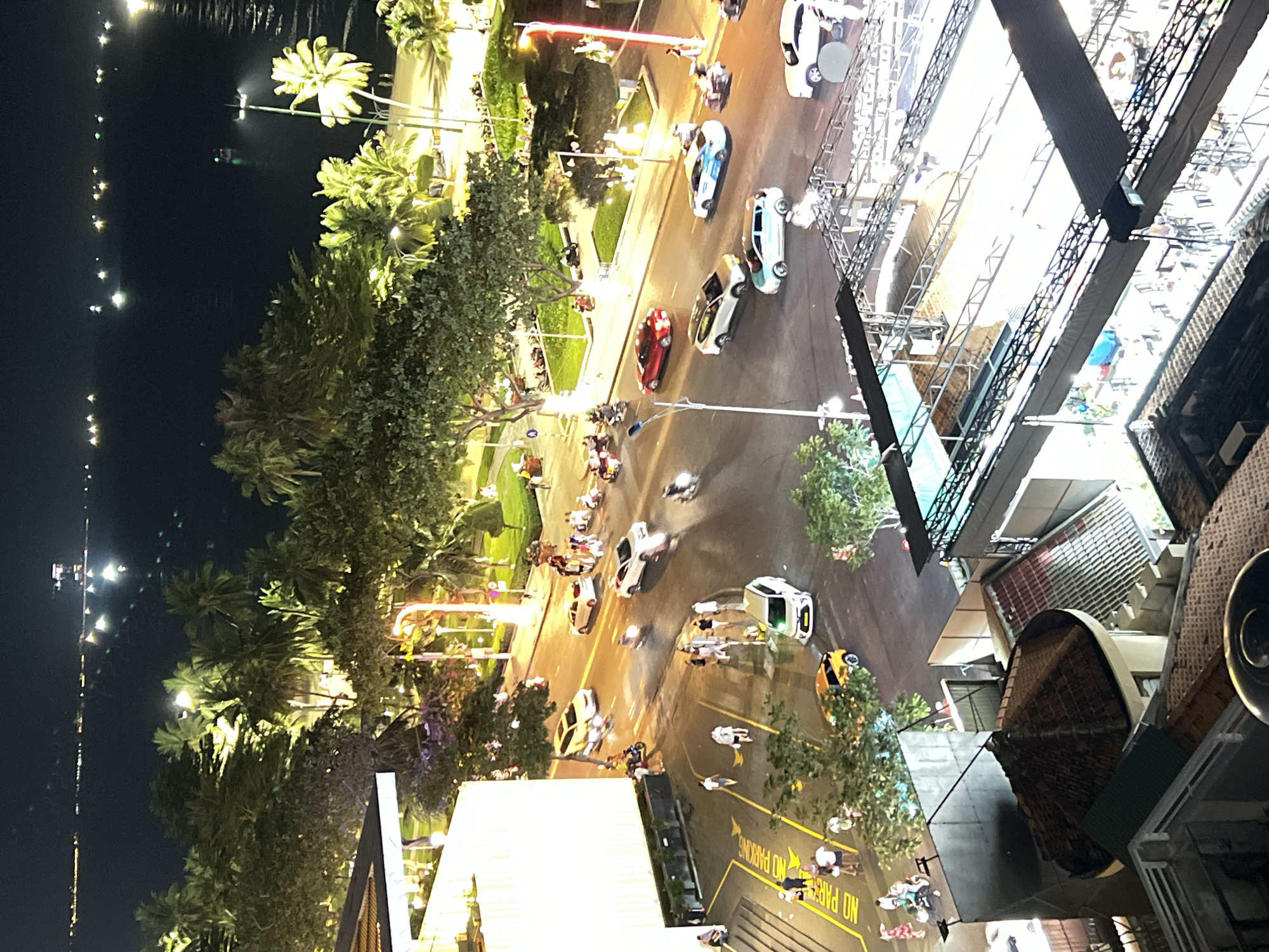 This is a nighttime aerial view of a street in Nha Trang, Vietnam, near the beach. The scene shows a busy street with various vehicles, including cars and motorcycles, driving and parked. Pedestrians are visible on the sidewalks and crossing the street. Palm trees and other greenery line the street, illuminated by streetlights. In the background, the ocean is visible with some lights from boats or structures on the water. The architecture suggests a coastal city with modern buildings and some older structures. The overall impression is a vibrant urban environment at night.