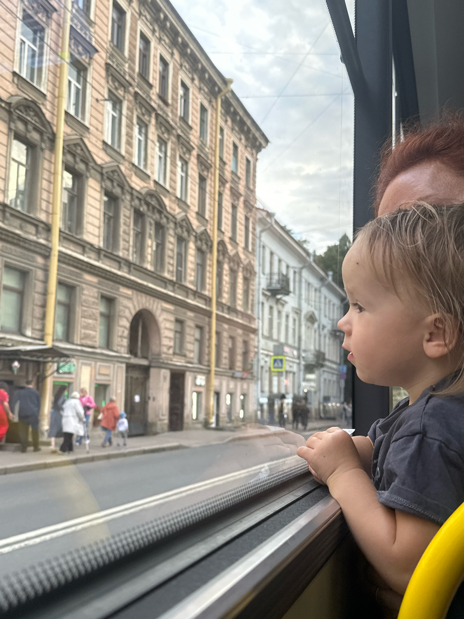 A young child with blond hair looks out a window, their hands resting on the sill. The child is wearing a dark t-shirt. To the left of the child, a person with red hair is partially visible. The window overlooks a city street in Saint Petersburg, Russia. Across the street are several multi-story buildings with classical architectural details. People are walking on the sidewalk in front of the buildings. The sky is overcast. The scene appears to be captured from inside a vehicle, likely a bus or tram, as indicated by the window frame and a yellow handrail.