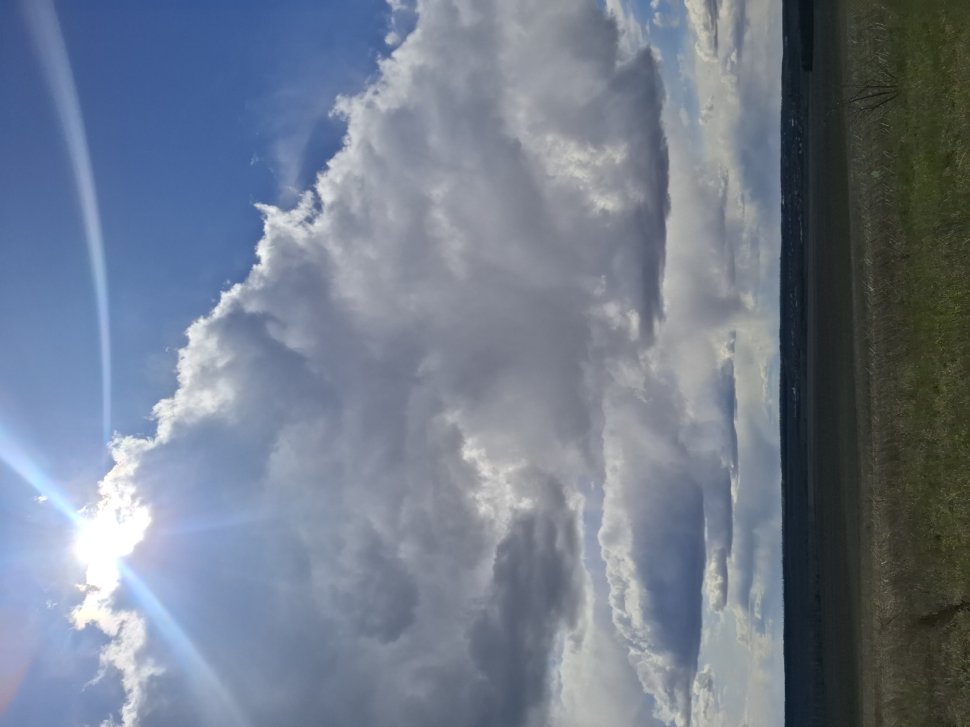 This media file presents a vast, open landscape under a dynamic sky, captured during daytime in the vicinity of Елец-Маланино, Russia. The sky dominates the scene, featuring a deep blue hue on the left that transitions into an expanse filled with large, voluminous cumulus clouds.
On the far left, the sun is partially hidden behind a cloud, producing a brilliant sunburst with prominent rays of light and a subtle lens flare arc in the upper left quadrant. The clouds display a variety of tones, from bright white where illuminated by the sun, to darker shades of gray in their shadowed depths, highlighting their texture and three-dimensionality.
Below the extensive sky, a horizontal strip of land stretches across the bottom of the image. This appears to be an open field or agricultural area, with visible patches of green vegetation and possibly some tilled earth. A distinct dark line across the horizon suggests a distant treeline, forest, or a remote settlement. A lone, sparsely branched tree or shrub is faintly discernible on the right edge of the field. No people, specific objects beyond the natural elements, or visible text are present in the image. The scene conveys a sense of expansive natural beauty on a bright, yet cloudy, day.