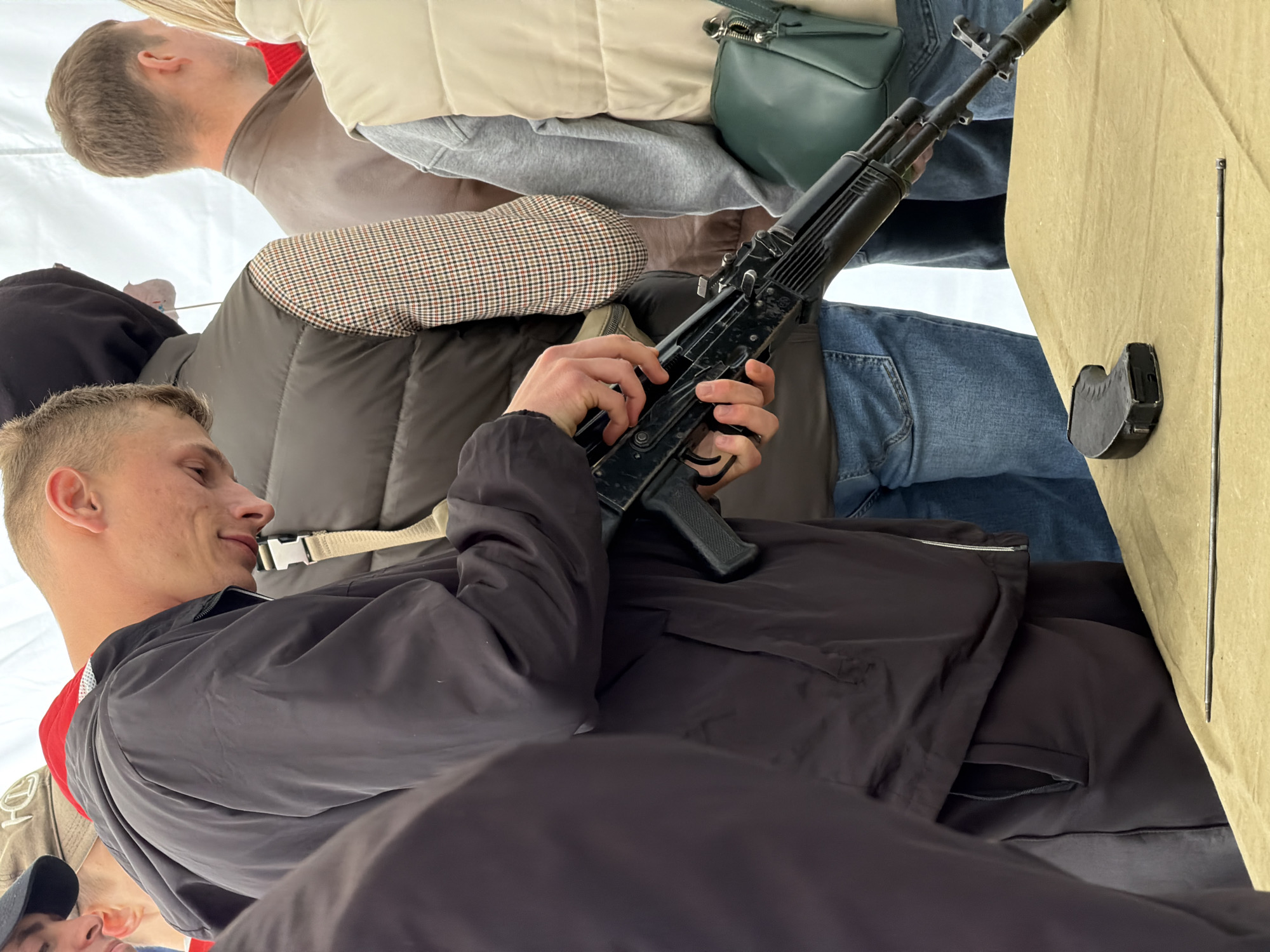 A medium shot taken indoors shows a man holding an AK-pattern rifle. The man is in the foreground, wearing a black jacket. He has short blonde hair and is looking to the left. The rifle is held in his hands, pointed upward. In the background are several other people, including one wearing a houndstooth-patterned jacket, another in jeans and a tan jacket with a green handbag, and another wearing a red top and ball cap. The photograph may have been taken from a low angle. A beige cloth or surface is visible to the right of the rifle. The location of Izhevsk, Russia is not evident from the photograph.