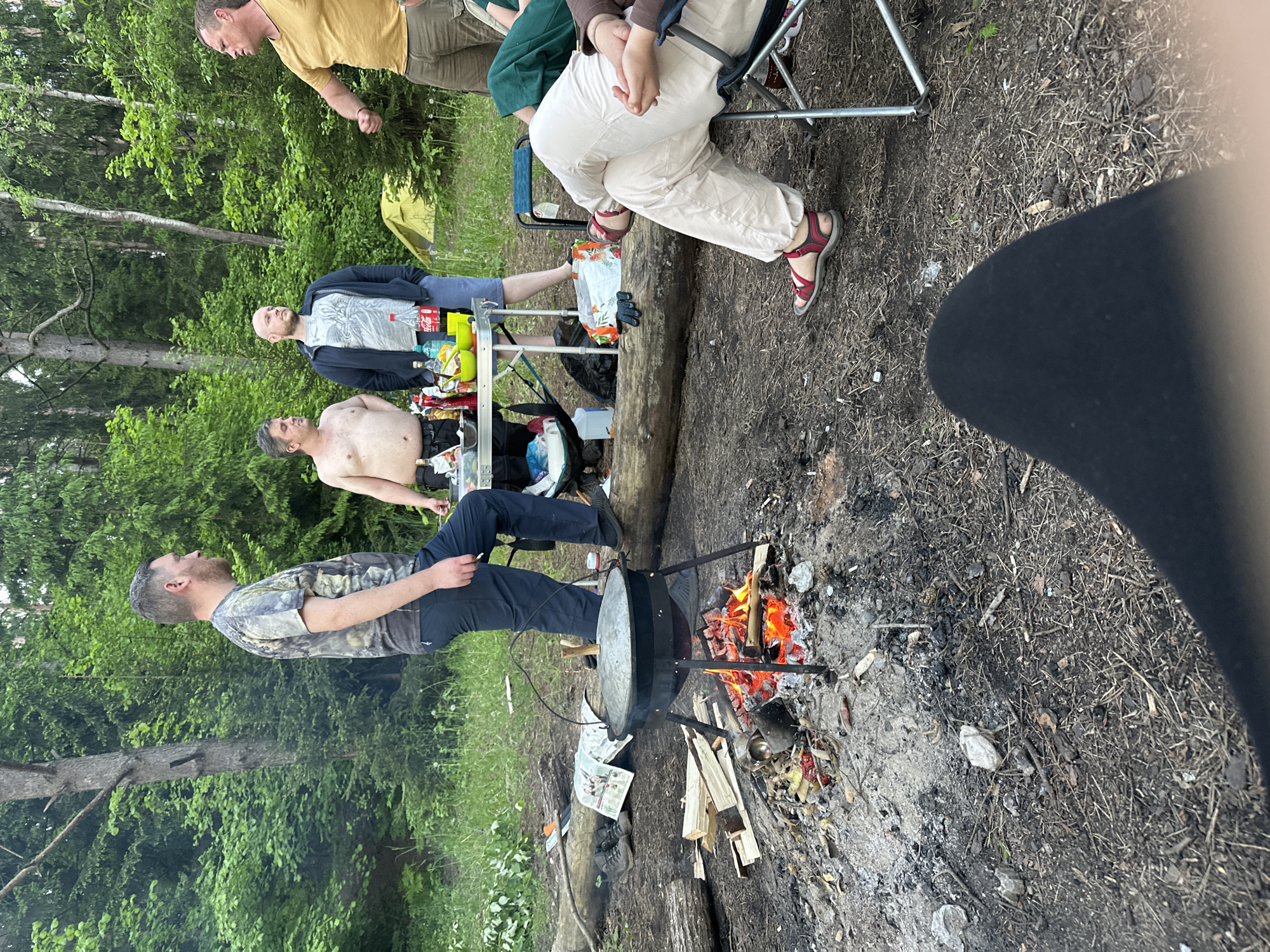The image depicts an outdoor scene in what appears to be a forested area near Юзуфоўскі сельскі Савет, Belarus. A group of people are gathered around a campfire, where a large cooking pot is suspended over the flames. A man in a camouflage shirt and dark pants stands near the fire. Another shirtless man is nearby near a table, along with a man wearing a gray t-shirt and jacket. Two other people are seated, one in green and the other in beige. Camping equipment, including a table, chairs, food, and other supplies, is scattered around the area. The environment is a typical forest setting with trees, grass, and a dirt ground covered with leaves and twigs.
