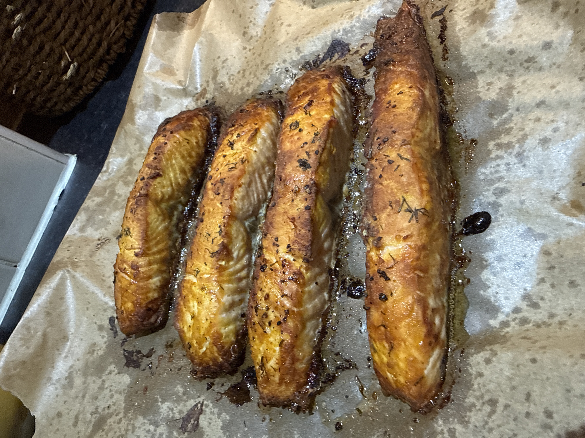 Four golden-brown pieces of cooked fish, likely salmon fillets, are arranged diagonally on a sheet of crinkled, grease-stained baking paper. The fish are glistening and show a slightly crispy, seasoned surface with visible dark specks, possibly herbs and spices like dried thyme or pepper. The fillets are irregularly shaped, suggesting they are natural cuts.
The baking paper beneath the fish is discolored by the cooking process, with dark, burnt residue and shimmering puddles of oil or fat, indicating the fish has just been cooked or is still warm. The paper is slightly rumpled and extends across most of the frame.
In the blurred background to the top left, a dark, woven basket-like object is partially visible, hinting at an indoor setting, likely a kitchen. Below it, to the far left, a corner of a white or light grey object, possibly a kitchen appliance or counter edge, is also present but out of focus. The lighting appears to be artificial, typical of an indoor kitchen environment. There is no visible text in the image. The scene captures a close-up of a prepared meal component. The specific location of Great Yarmouth, United Kingdom, cannot be discerned from the visual information provided.